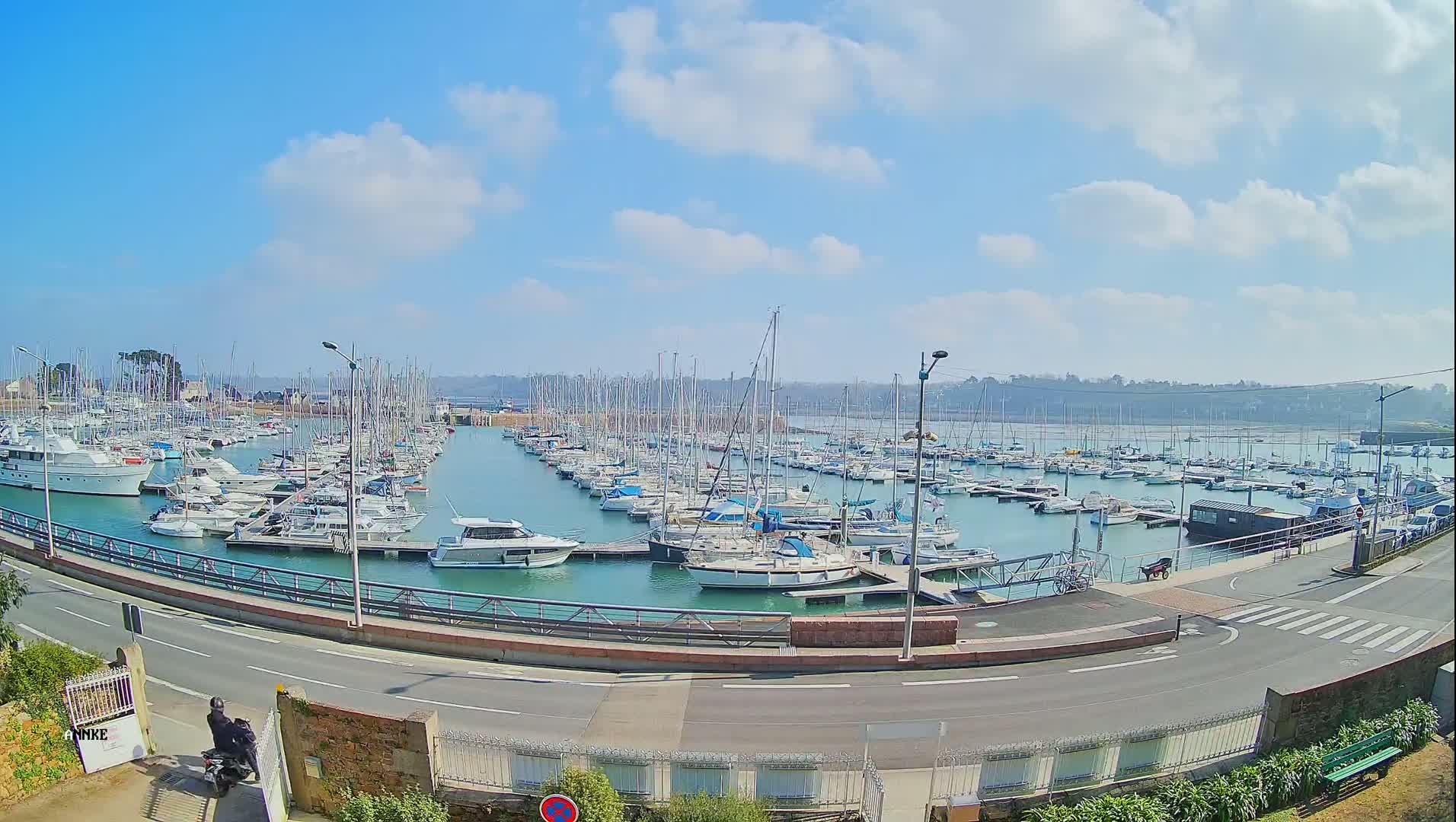 A marina filled with numerous sailboats and motorboats is seen on a sunny day with scattered clouds.