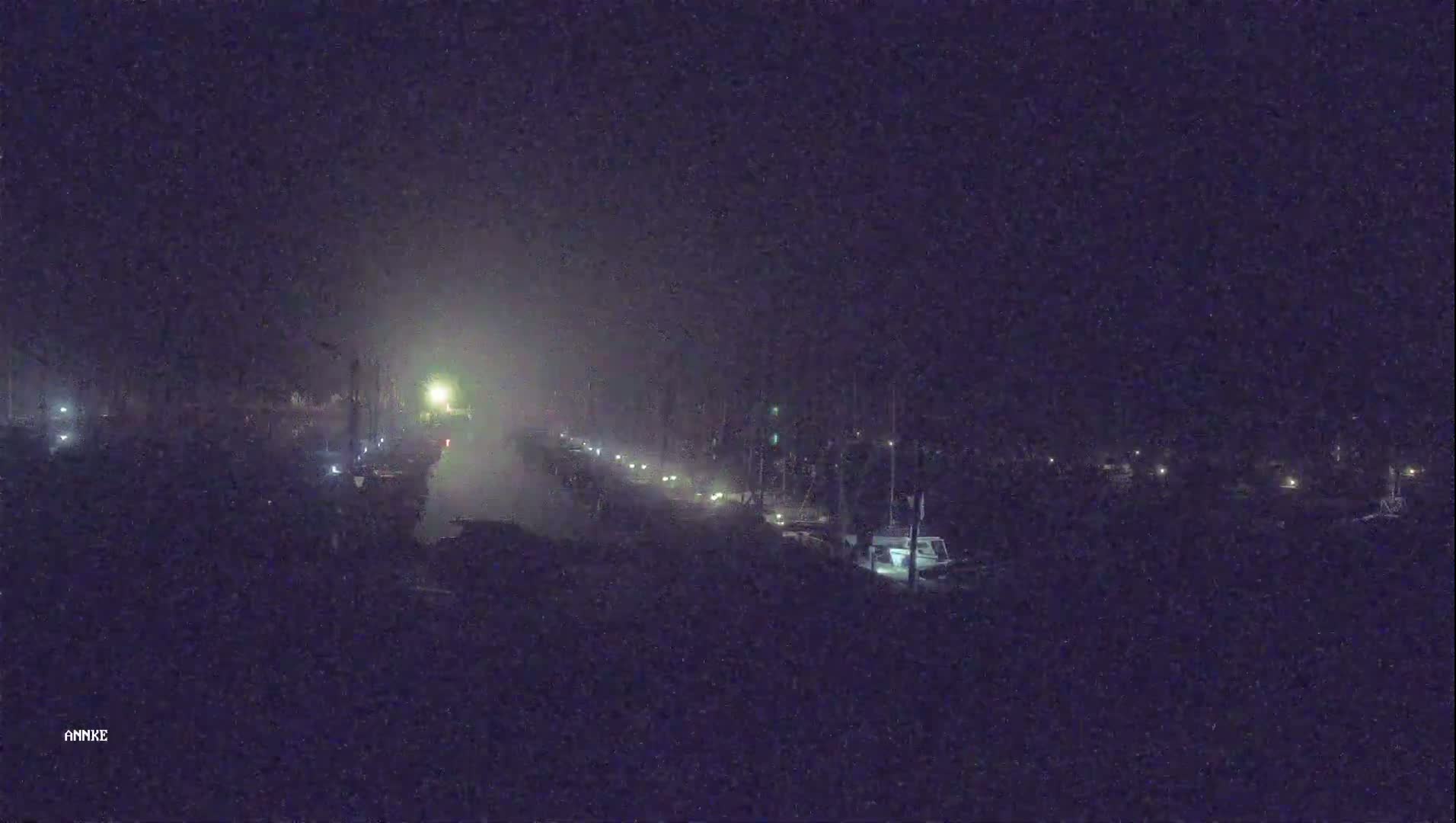 A foggy night scene shows a dimly lit waterway with boats moored on either side, and a few lights visible along a road or path in the center.