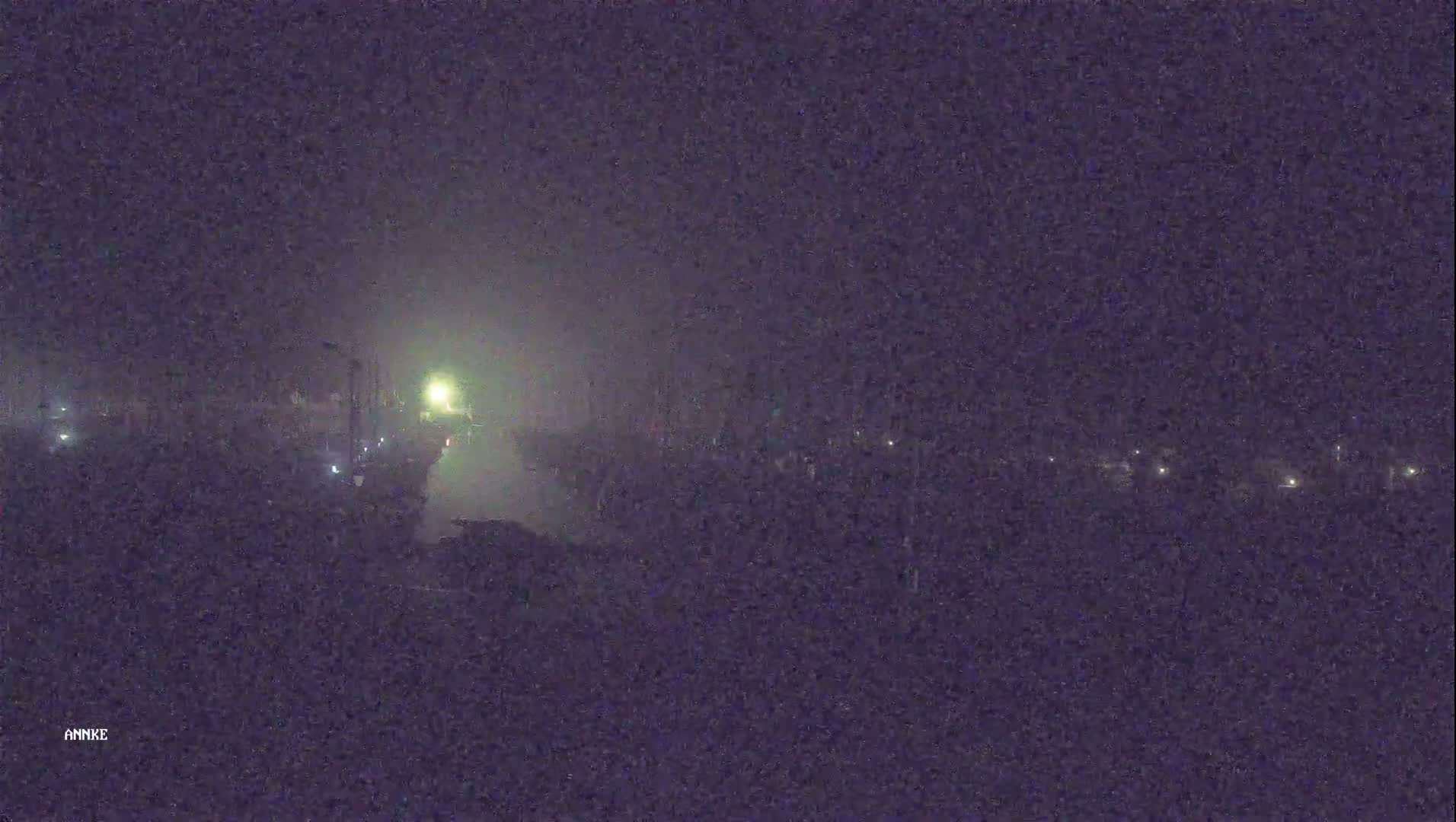 A nighttime, foggy scene shows a dimly lit roadway with a few distant lights visible.
