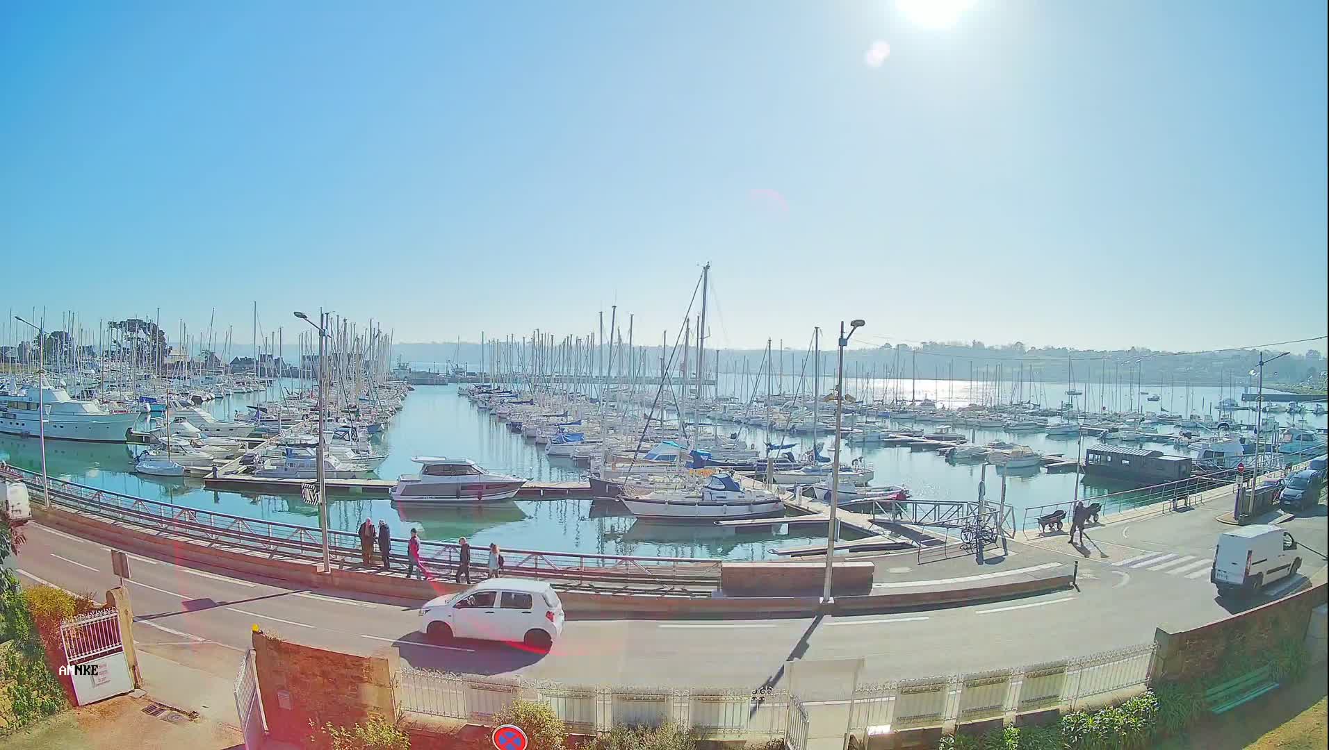 A sunny day at a marina filled with numerous sailboats and motorboats, with a few people walking along a road next to the water.