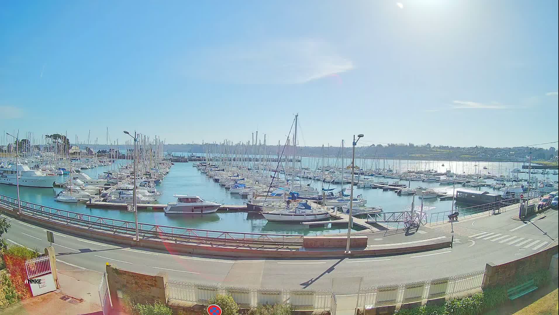 A sunny day at a marina filled with numerous sailboats and motorboats, a road curves around the harbor.