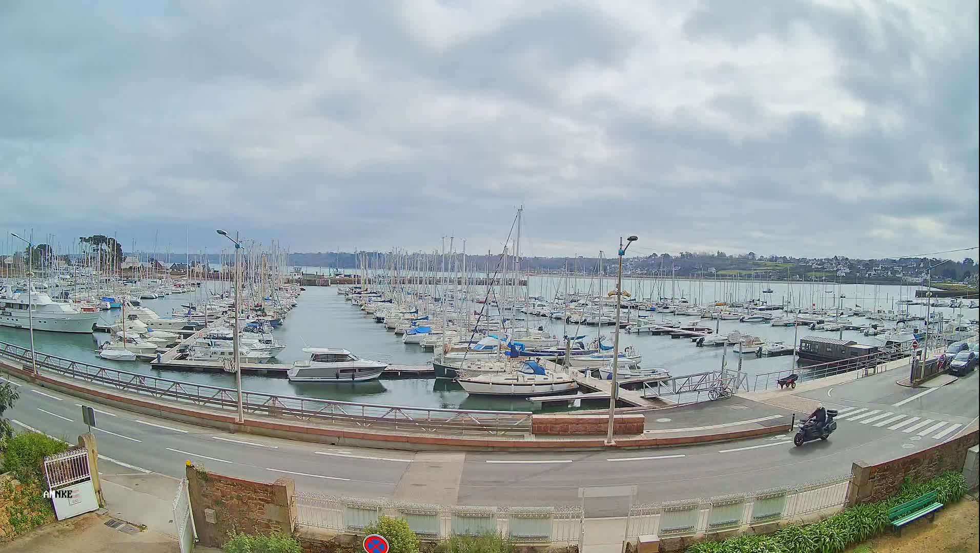 A large marina filled with numerous sailboats and motorboats is seen under an overcast sky, with a road and a person on a motorcycle visible in the foreground.