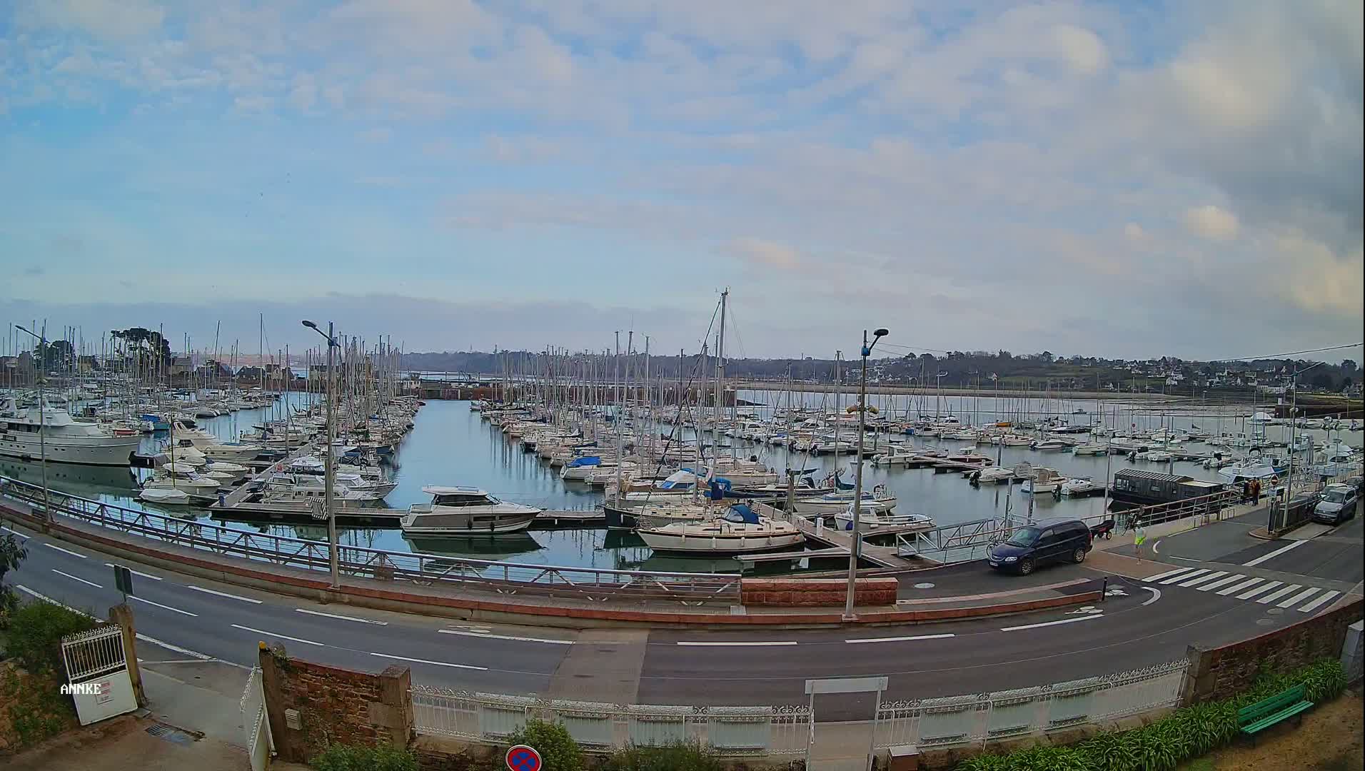 A marina filled with numerous sailboats and motorboats is viewed from across a road under a partly cloudy sky.