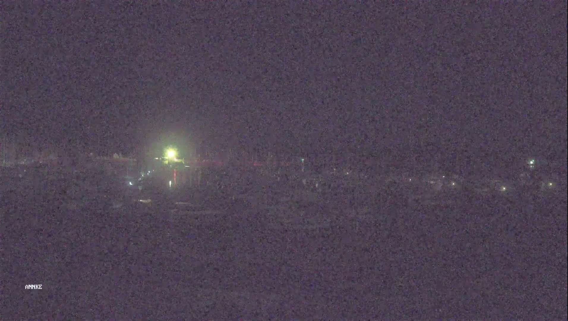 A nighttime, low-light view shows a dark landscape with a few scattered lights in the distance, possibly a body of water reflecting some light.
