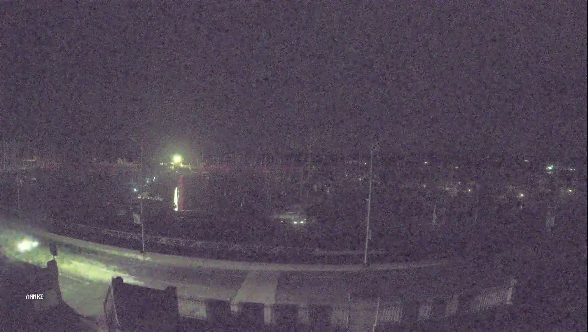 A nighttime, high-angle view shows a dark road curving through a parking lot and residential area with sparsely visible vehicles and lights.