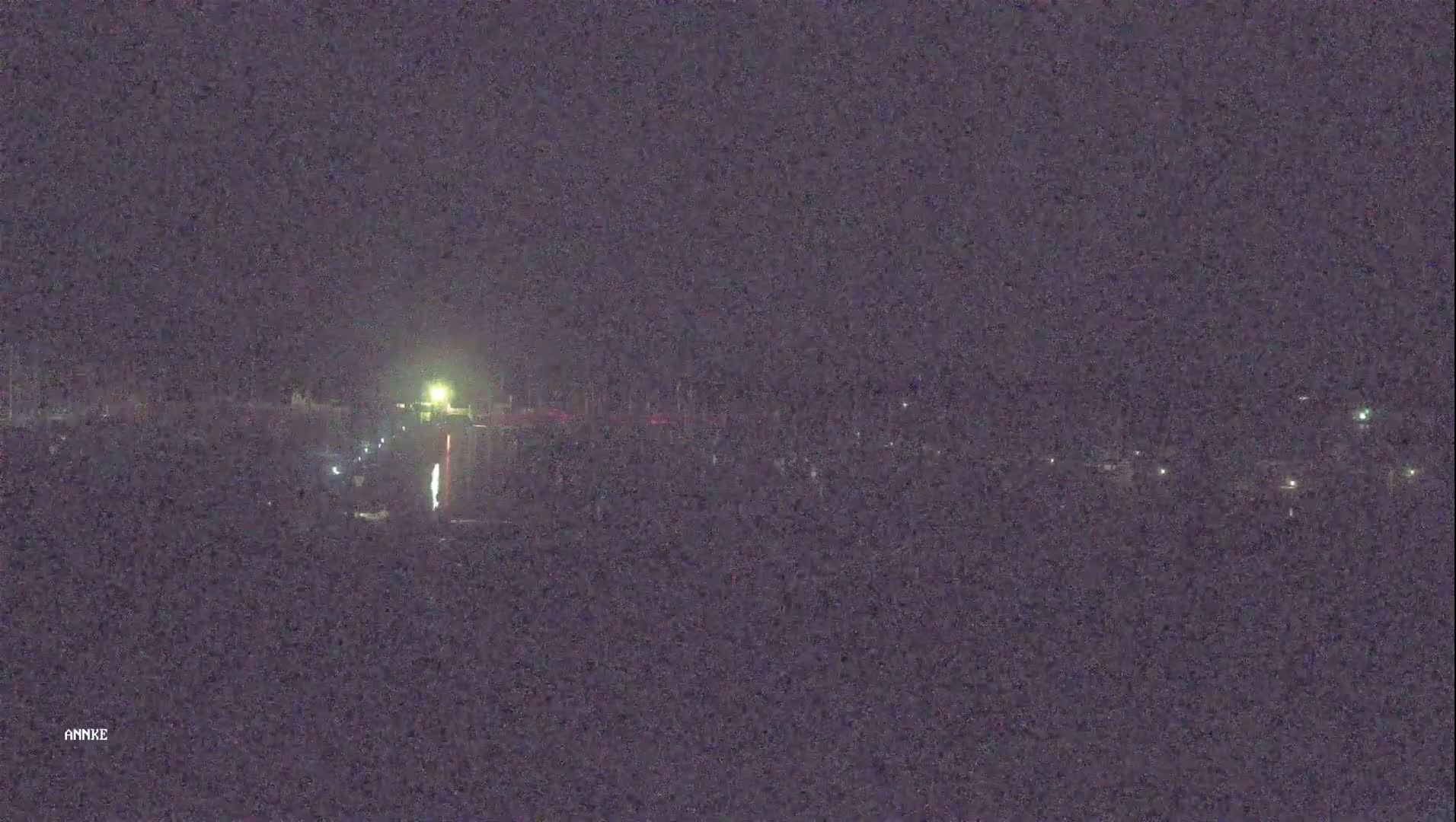 A nighttime long shot shows a dimly lit area with scattered lights, possibly a parking lot or a waterfront, under a dark sky.
