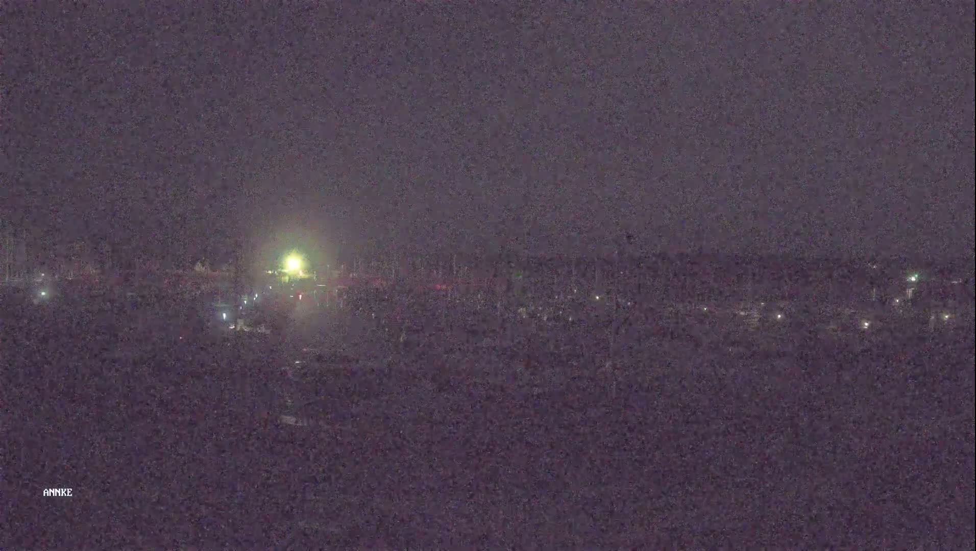 A nighttime, long shot of a dimly lit area with scattered lights, possibly a parking lot or a residential area, under a dark sky.