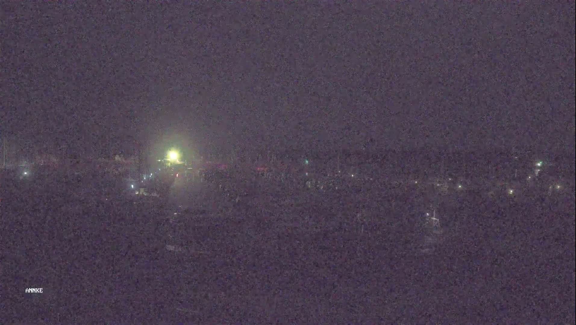 A nighttime, long shot shows a sparsely lit area with scattered lights and a brighter light source in the distance.
