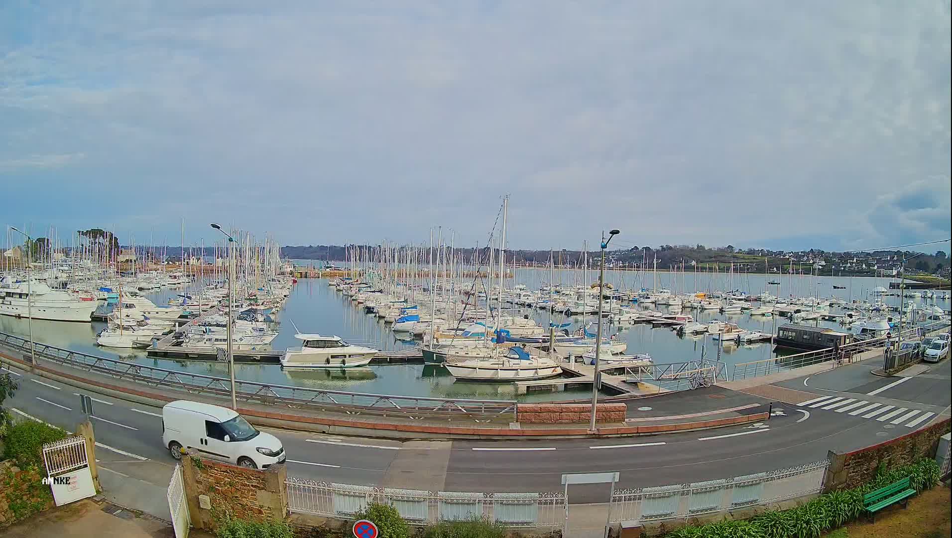A marina filled with numerous sailboats and motorboats under a mostly cloudy sky, with a white van driving on a road next to it.