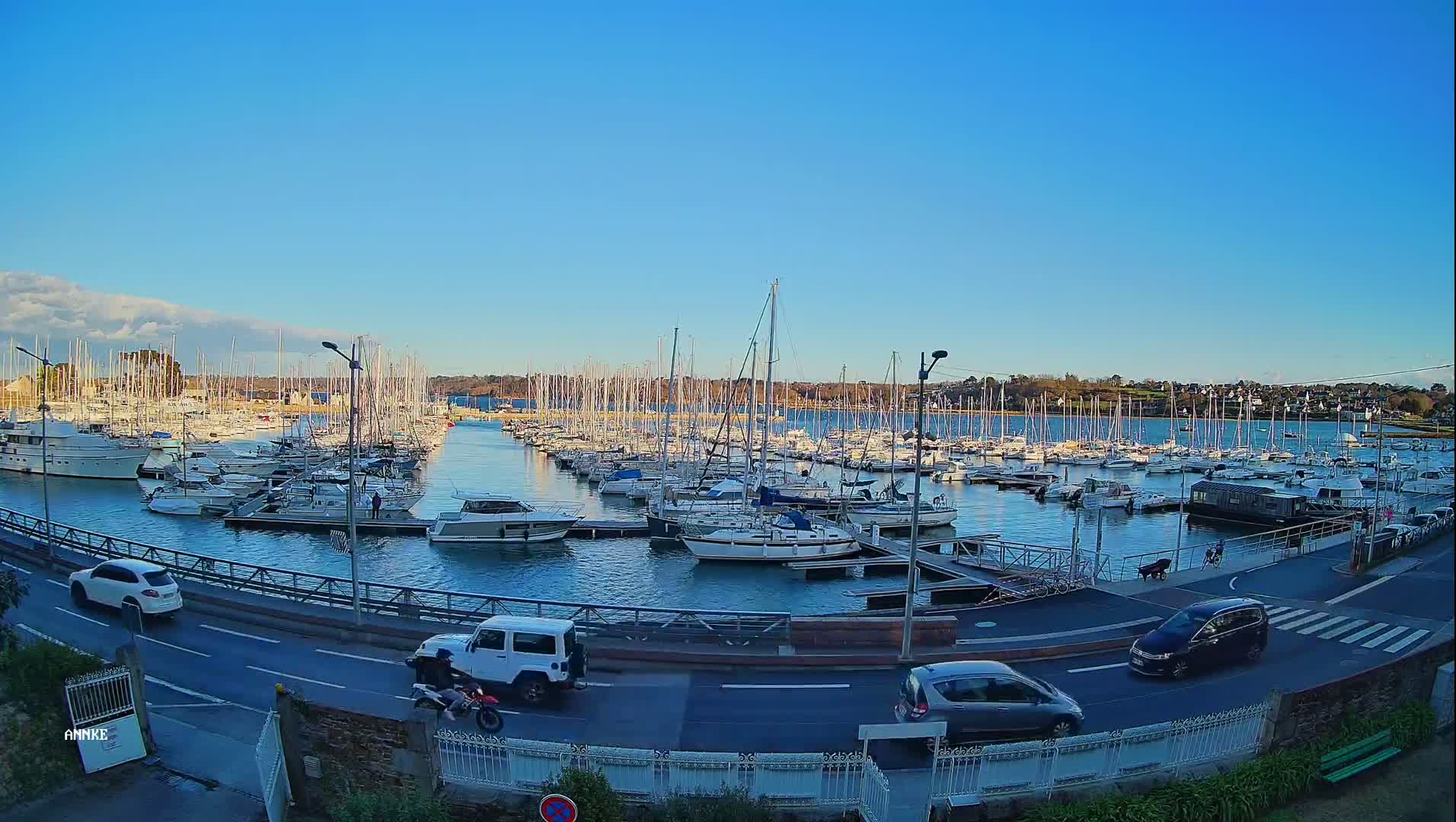 A marina filled with sailboats and motorboats is viewed from above on a clear, sunny day, with cars driving on a road next to the water.
