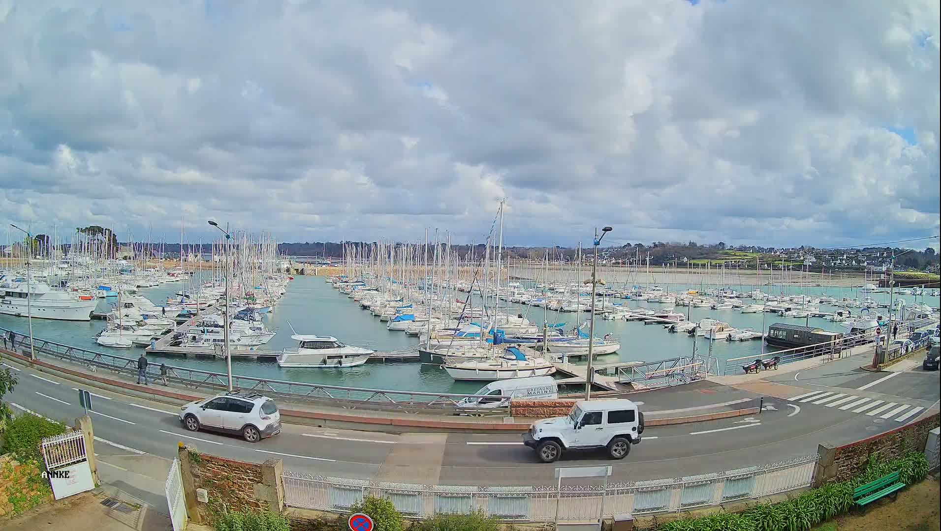 A large marina filled with numerous boats is viewed from above on a partly cloudy day, with cars driving along a road next to the water.