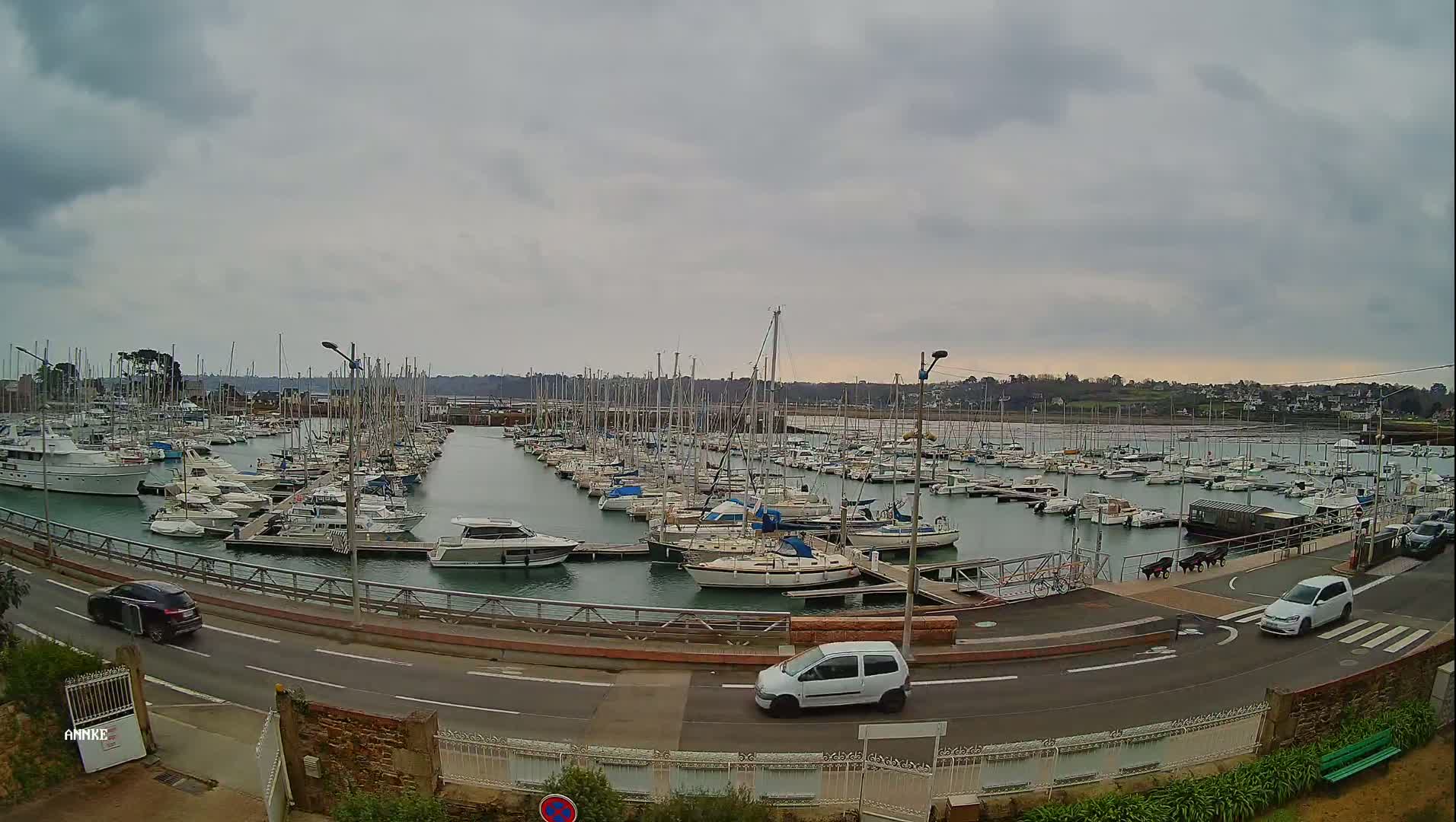 A marina filled with numerous sailboats and motorboats is seen under an overcast sky, with cars driving along a nearby road.