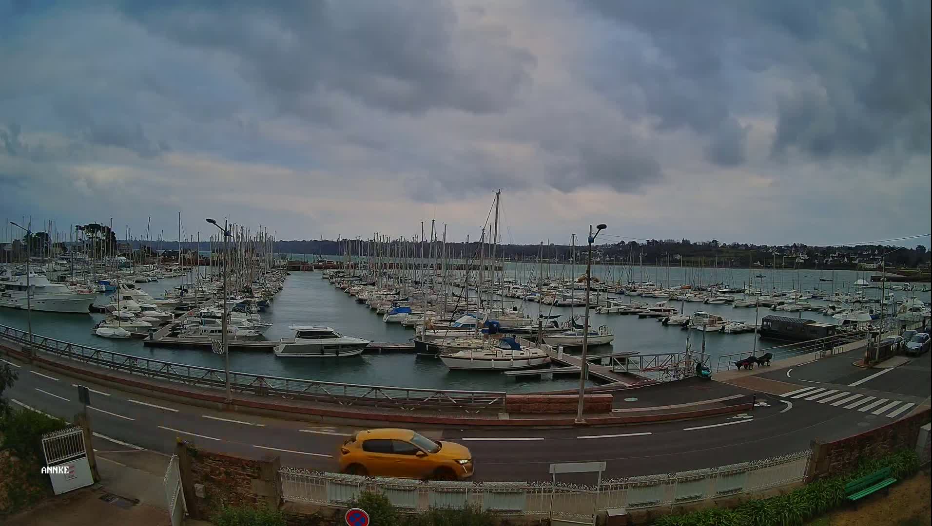 A large marina filled with numerous sailboats and motorboats is seen under a cloudy sky, with a yellow car driving along a road beside it.