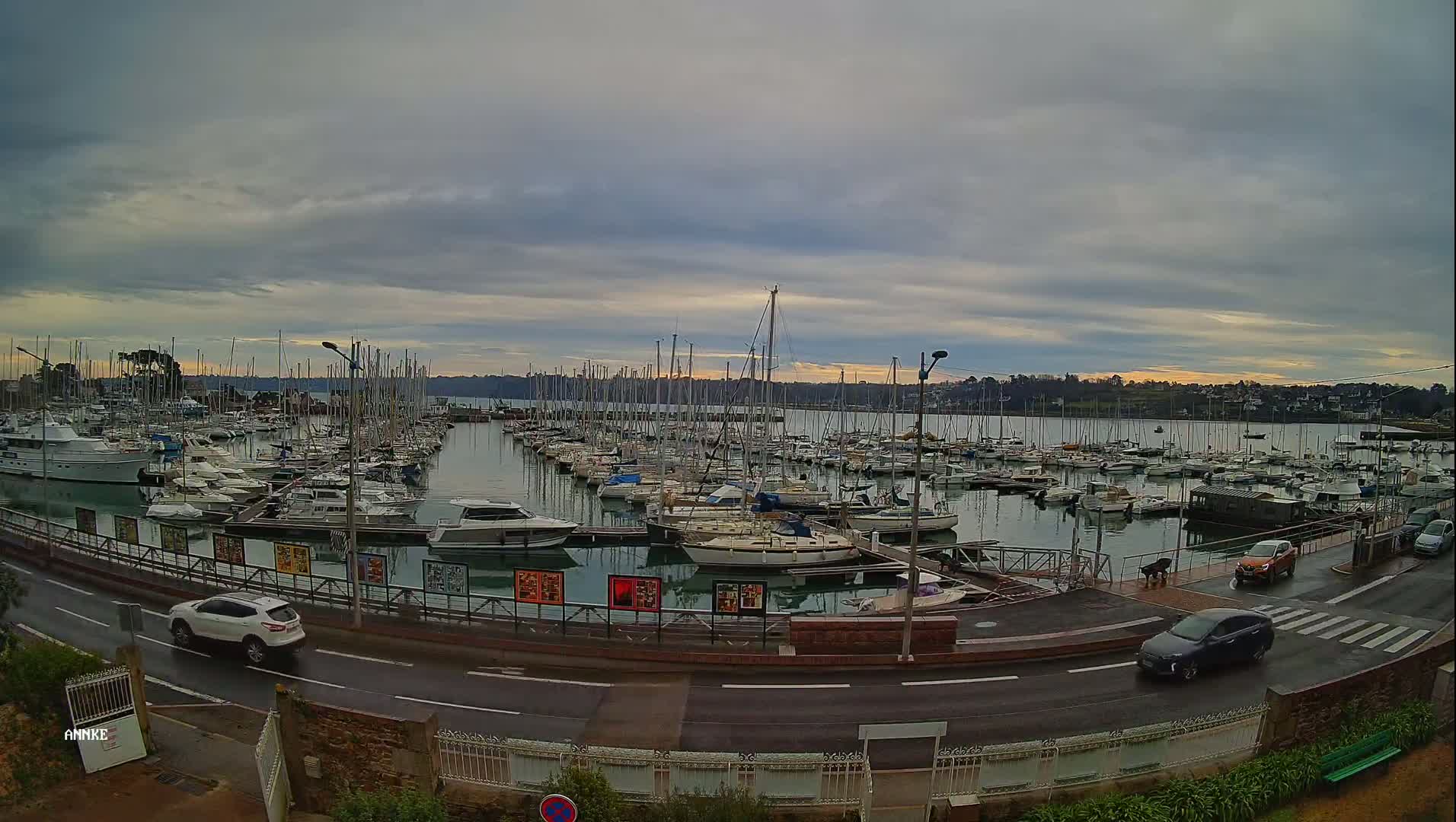 A marina filled with numerous sailboats and motorboats is seen under a cloudy sky, with cars driving along a road next to the water.