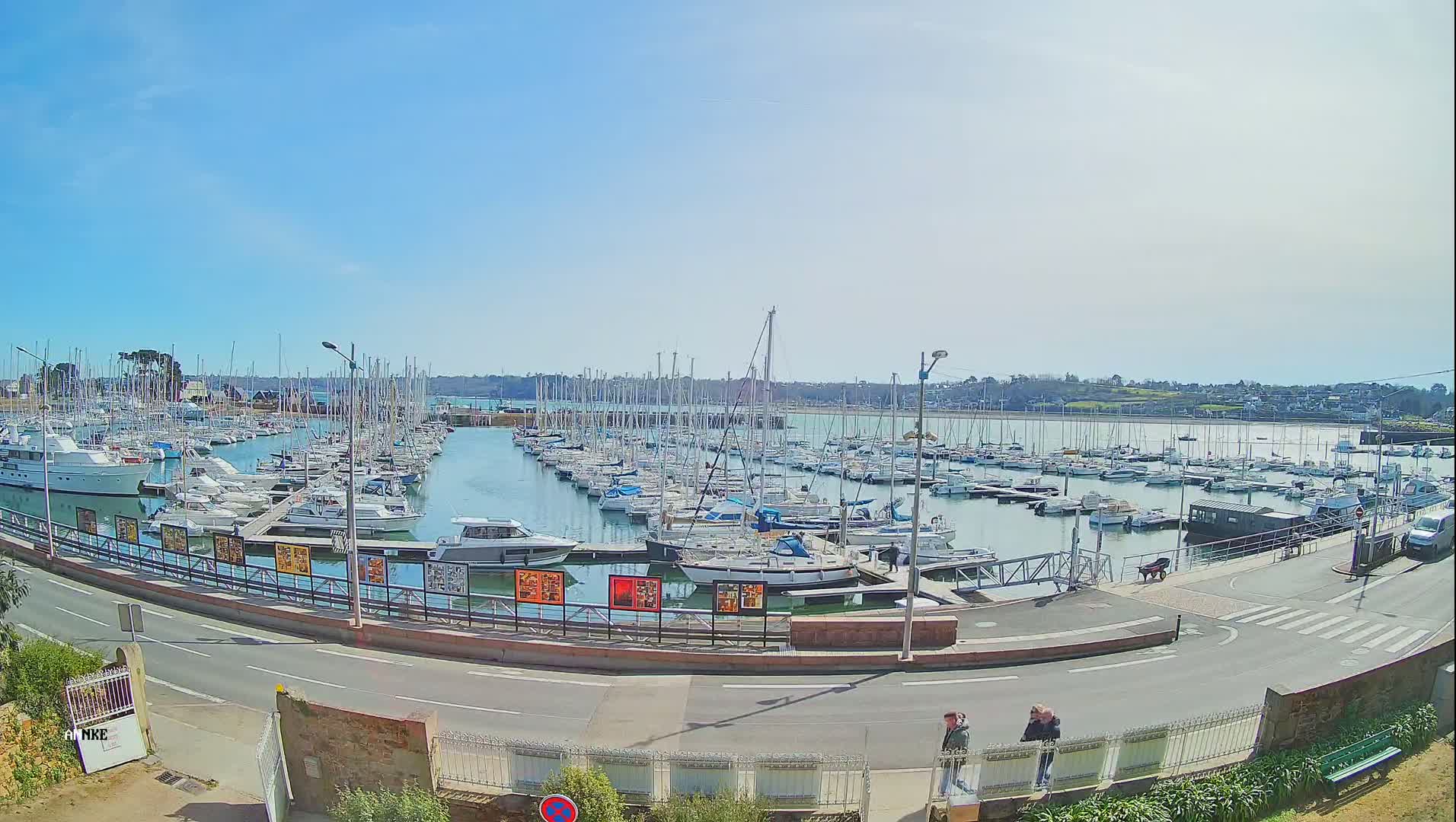 A sunny day shows a marina filled with numerous sailboats and motorboats, with a road and pedestrians visible in the foreground.