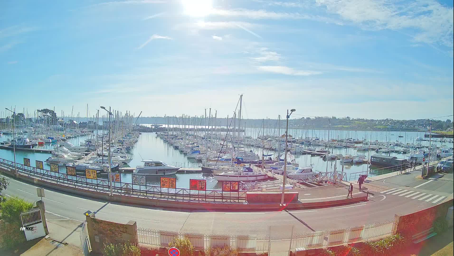 A sunny day at a marina shows numerous sailboats and motorboats docked, with a roadway curving around the harbor.