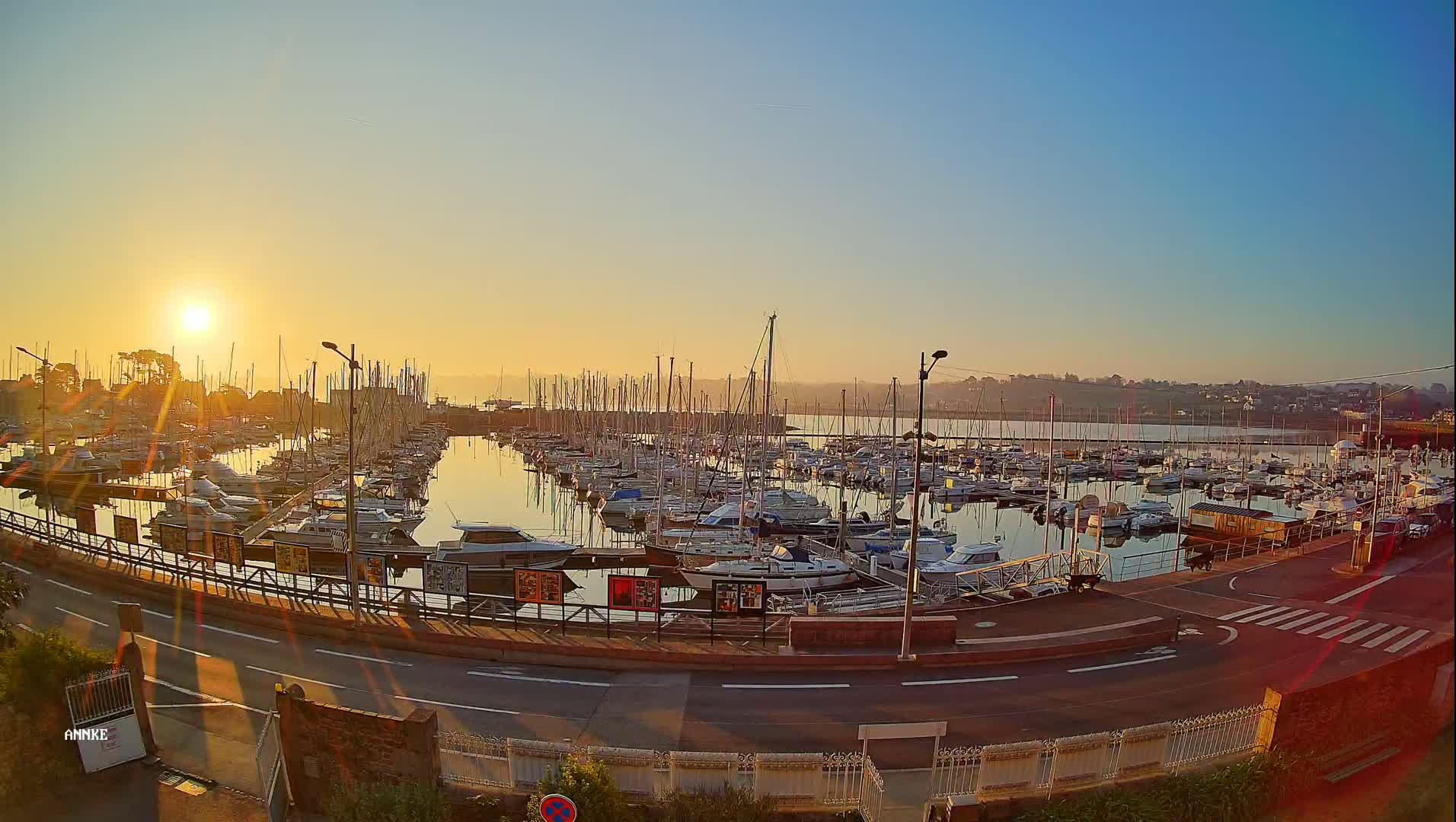 A marina filled with numerous sailboats and motorboats is bathed in the golden light of sunrise over calm water.