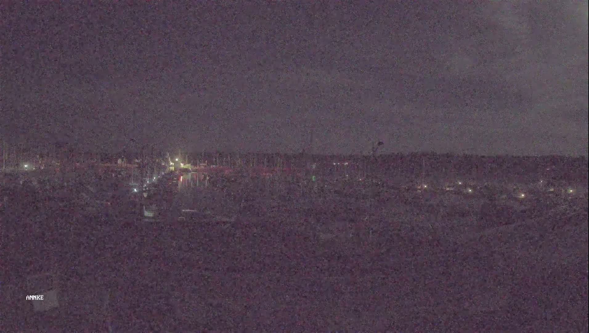 A nighttime long shot shows a dark body of water with numerous small lights along its edges, under a dark, cloudy sky.