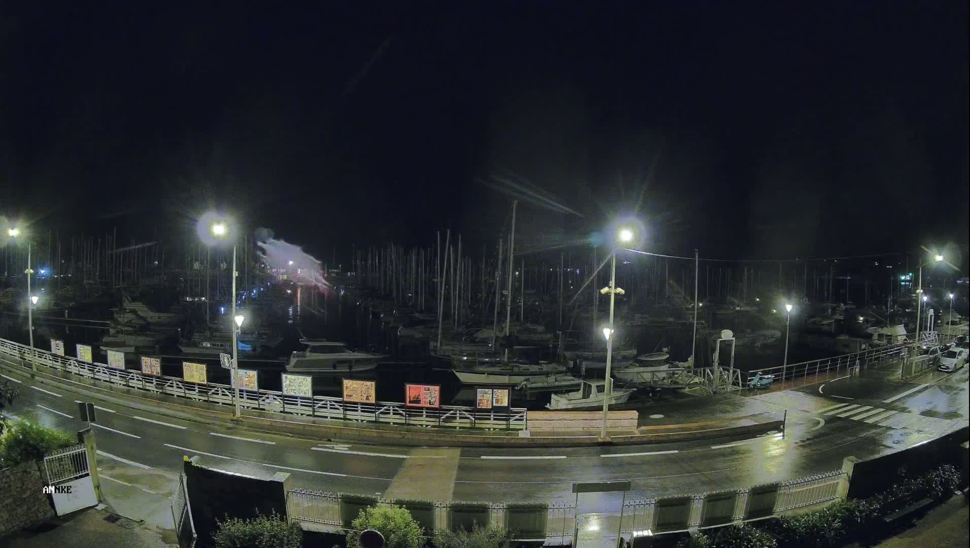 A wet, nighttime marina with many boats docked along a roadway under bright streetlights.