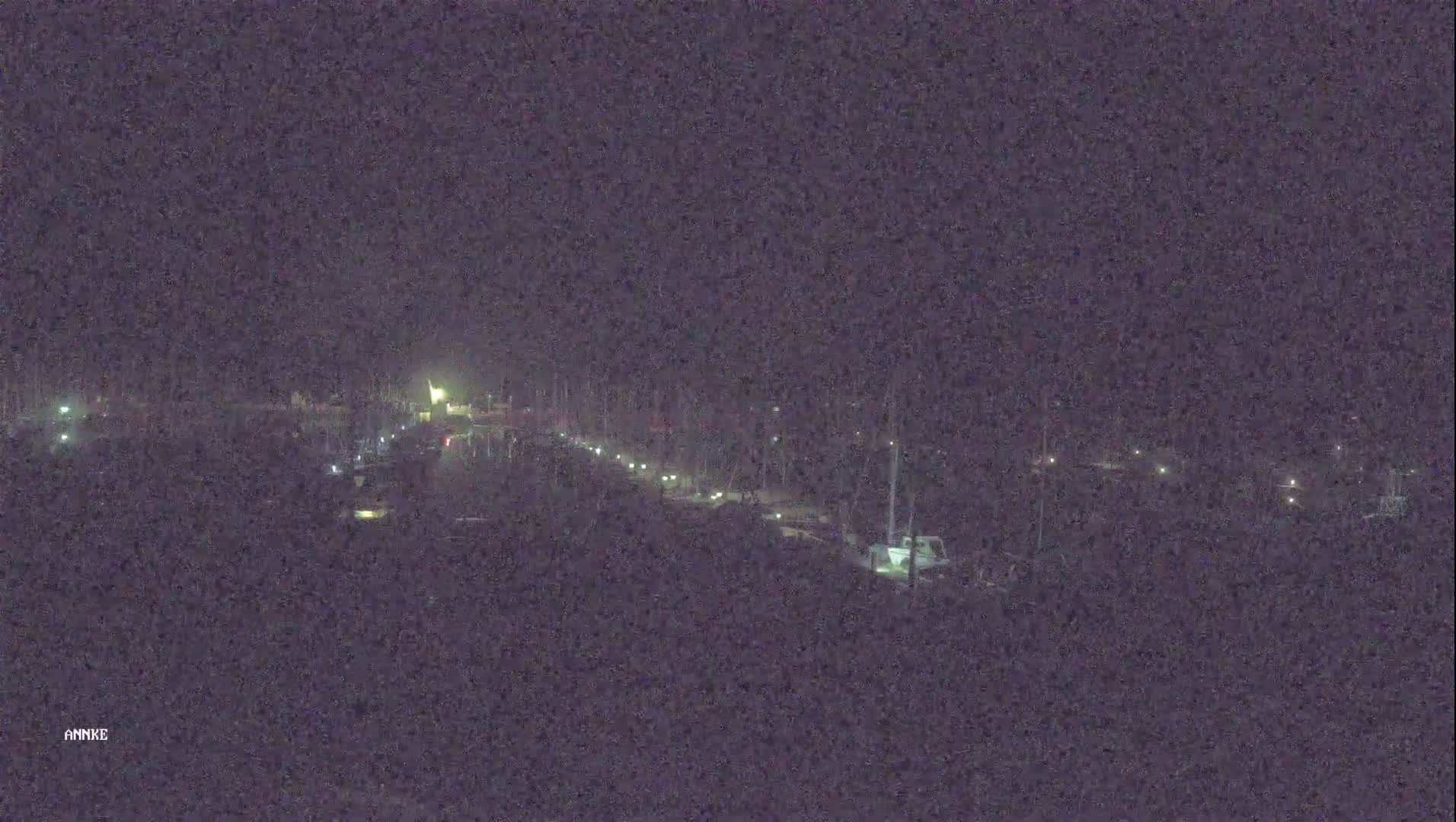 A nighttime long shot shows a road with sparsely spaced vehicles and dimly lit buildings or structures on either side.