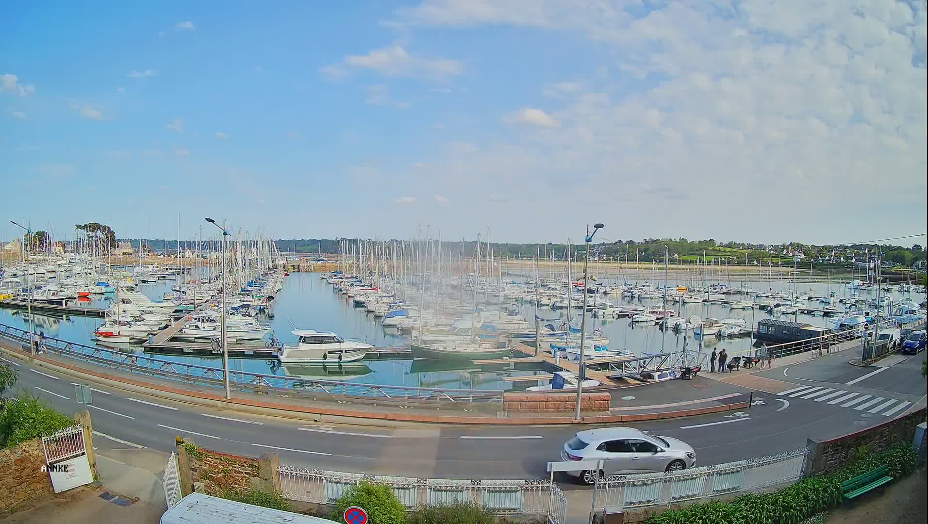 A marina filled with numerous sailboats and motorboats is seen from across a road on a partly cloudy day.