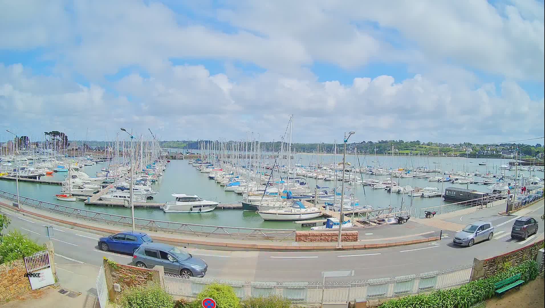 A marina filled with numerous sailboats and motorboats is seen under a partly cloudy sky, with cars parked along a road overlooking the water.