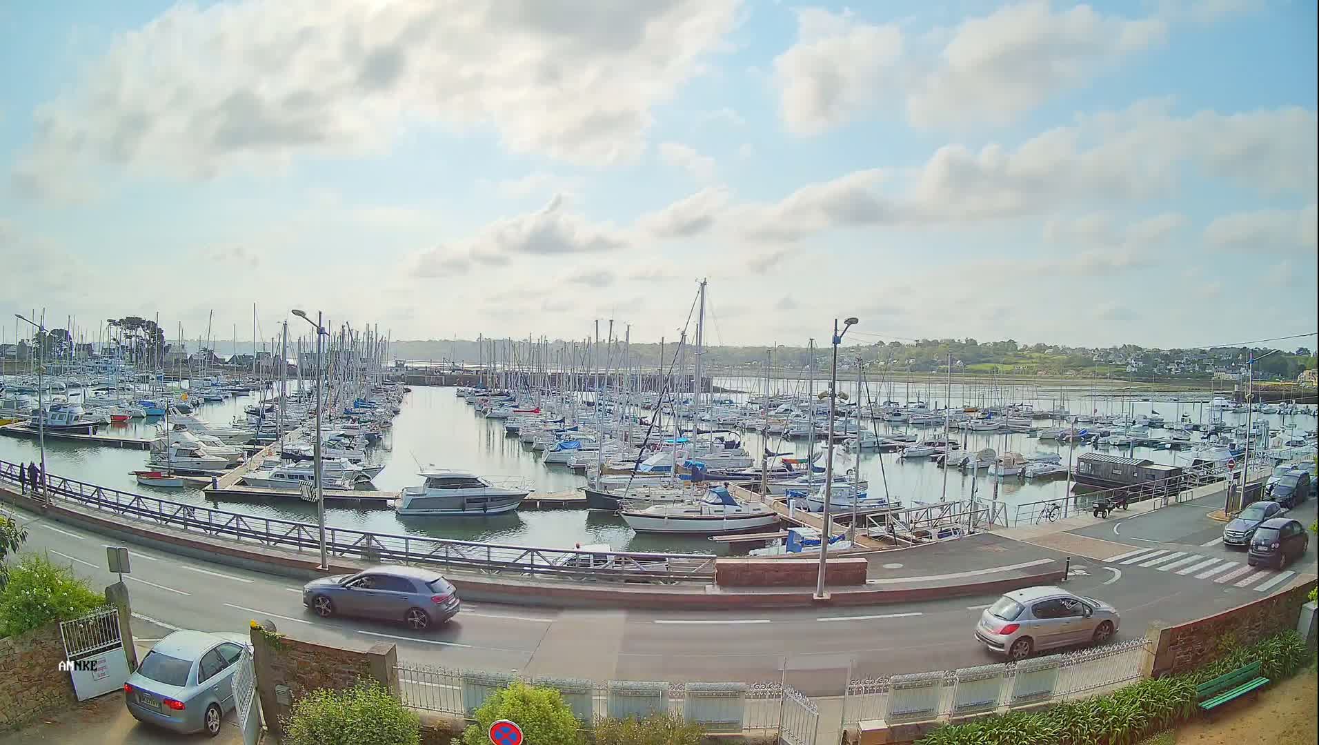 A marina filled with numerous sailboats and motorboats is situated next to a road with several cars parked and driving under a partly cloudy sky.