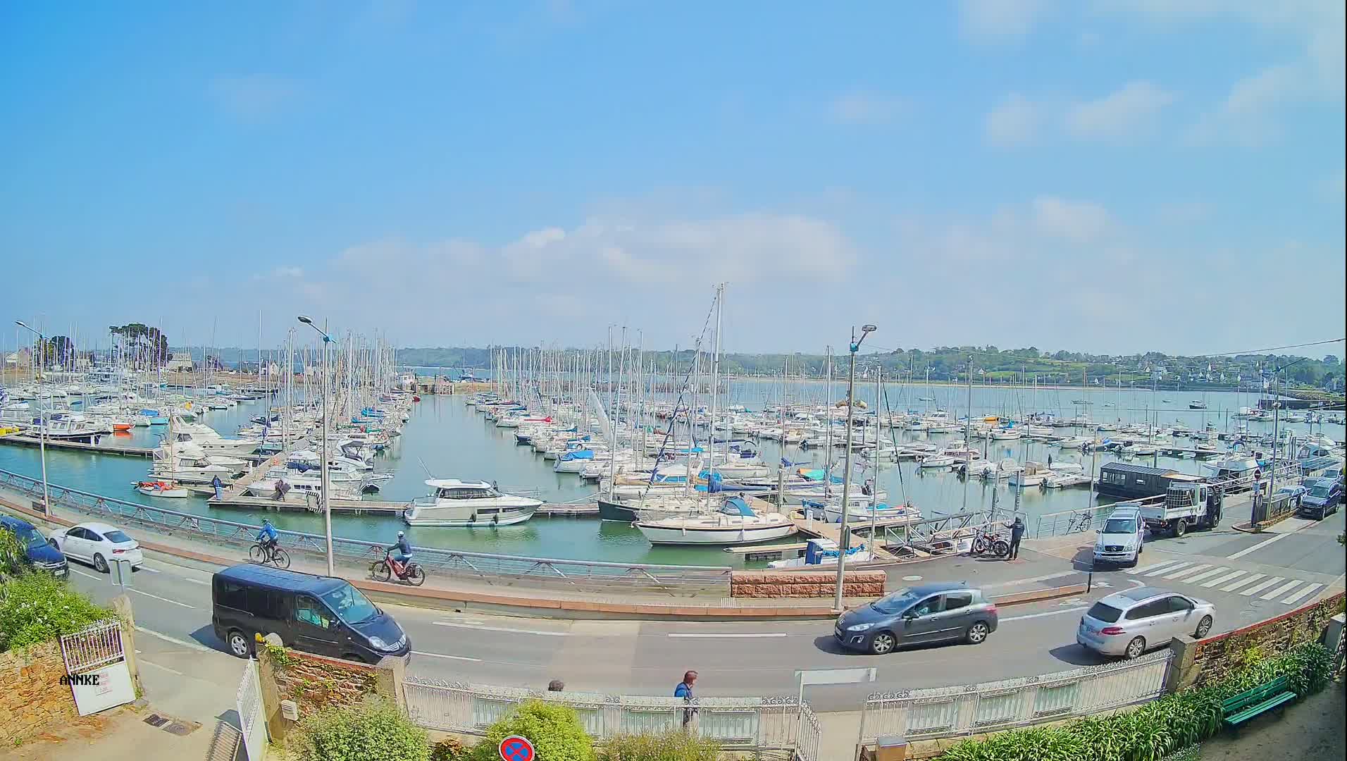 A sunny day shows a large marina filled with sailboats and motorboats, with cars and cyclists on a road nearby.