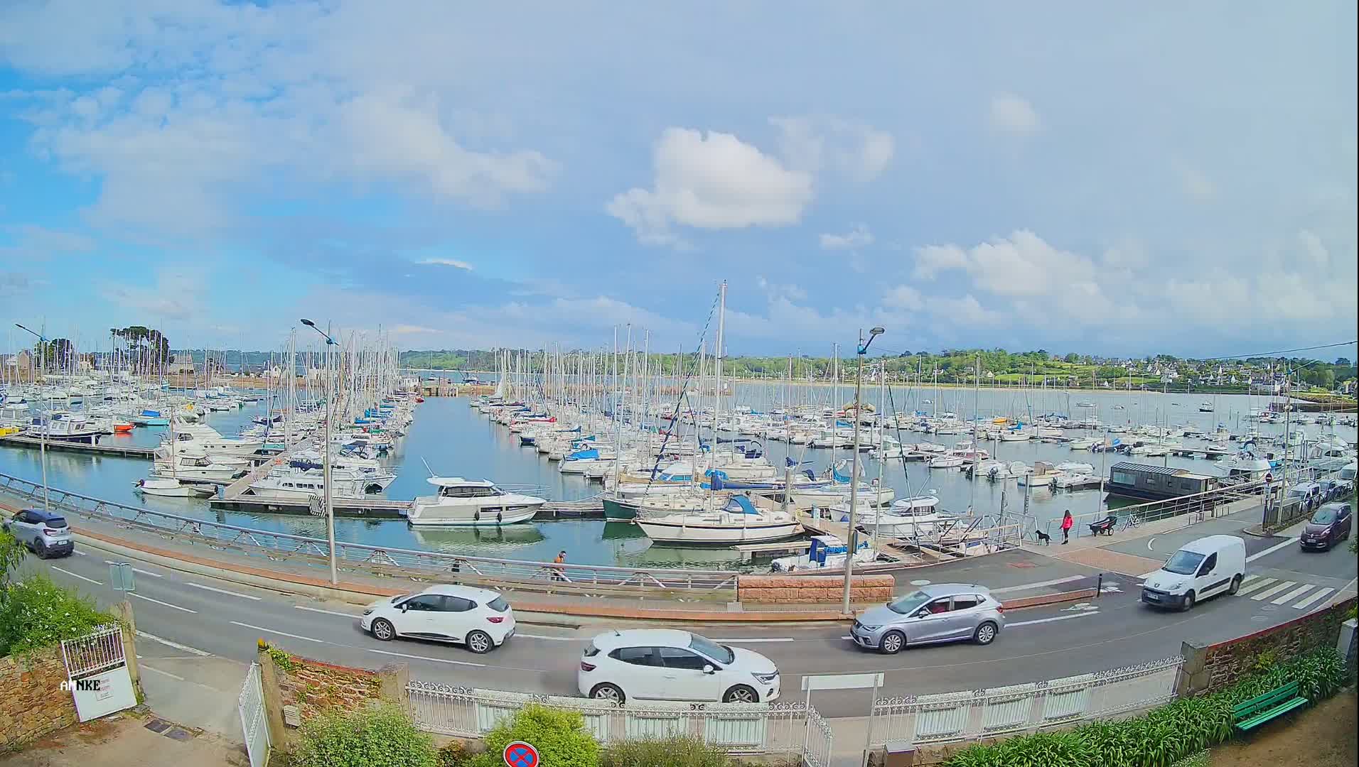 A large marina filled with sailboats and motorboats is viewed from a nearby road under a partly cloudy sky.