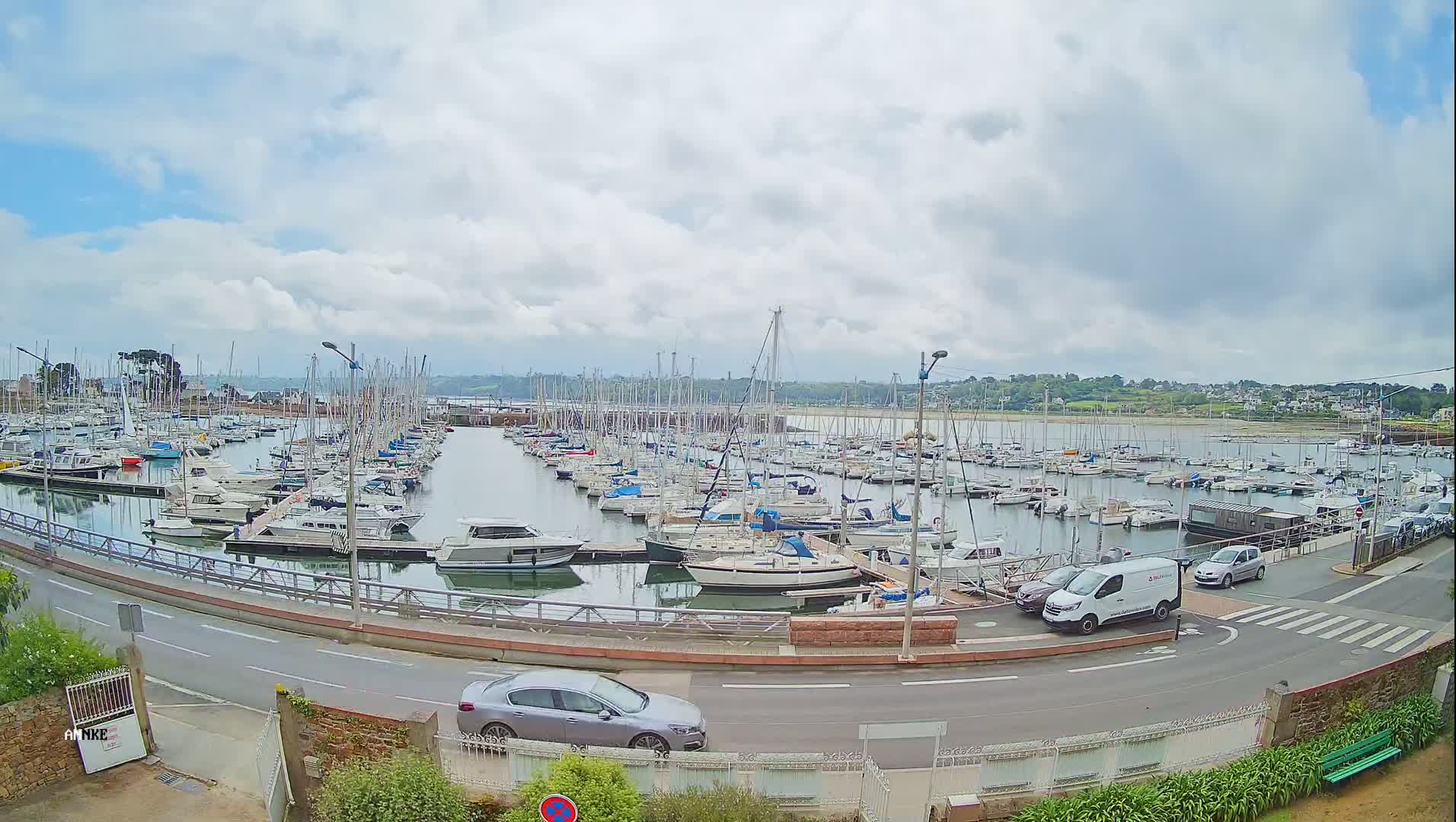 A harbor filled with numerous sailboats and motorboats under a partly cloudy sky, with a road and several vehicles visible in the foreground.