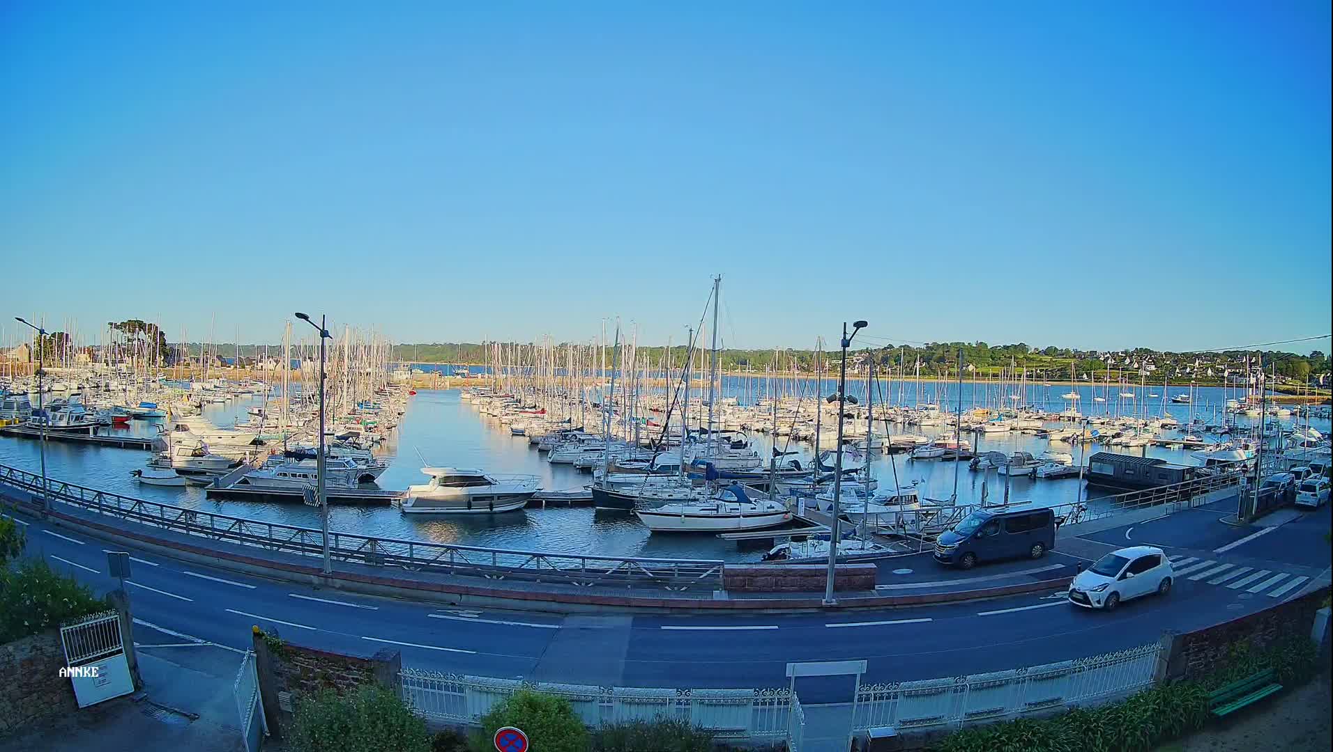 A marina filled with numerous sailboats and motorboats under a clear blue sky is viewed from above a road next to the water.