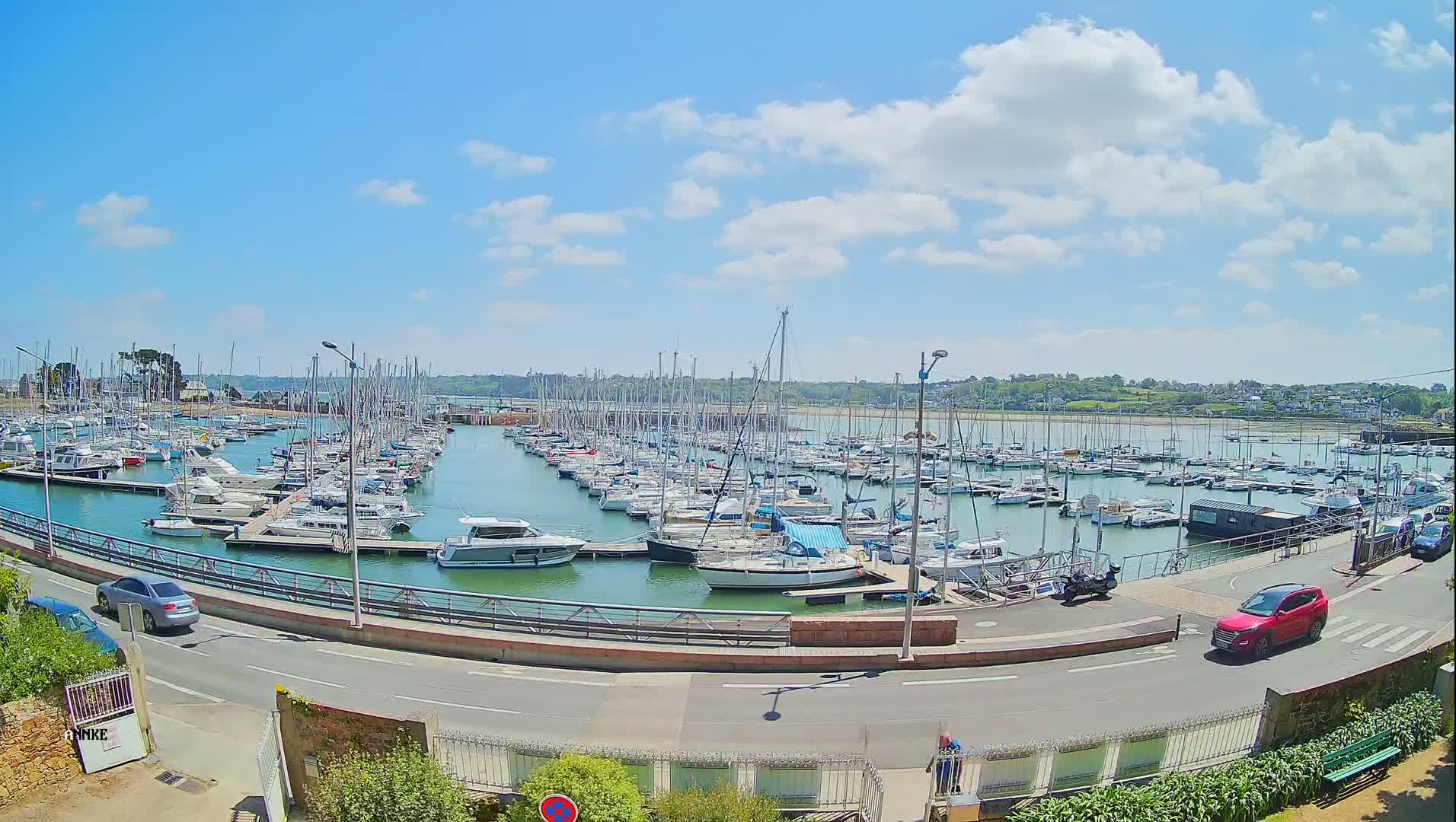 A marina filled with numerous sailboats and motorboats is situated next to a road on a sunny day with scattered clouds.