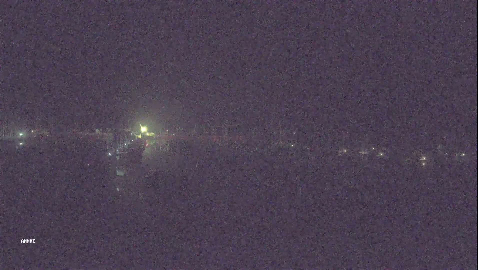 A nighttime, low-light view shows a long row of dimly lit objects, possibly vehicles, near a body of dark water under a dark sky.