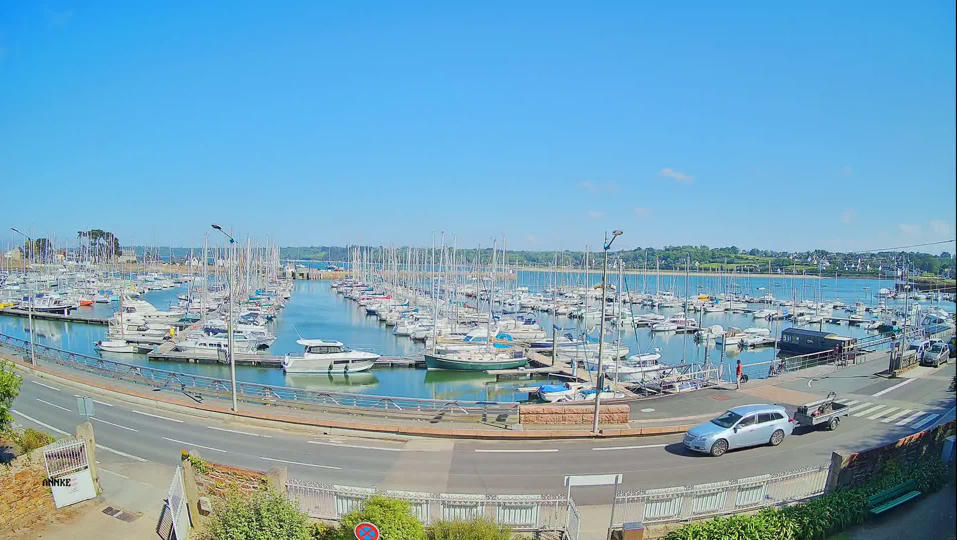 A marina filled with numerous sailboats and motorboats is situated on calm, clear water under a bright, sunny sky.