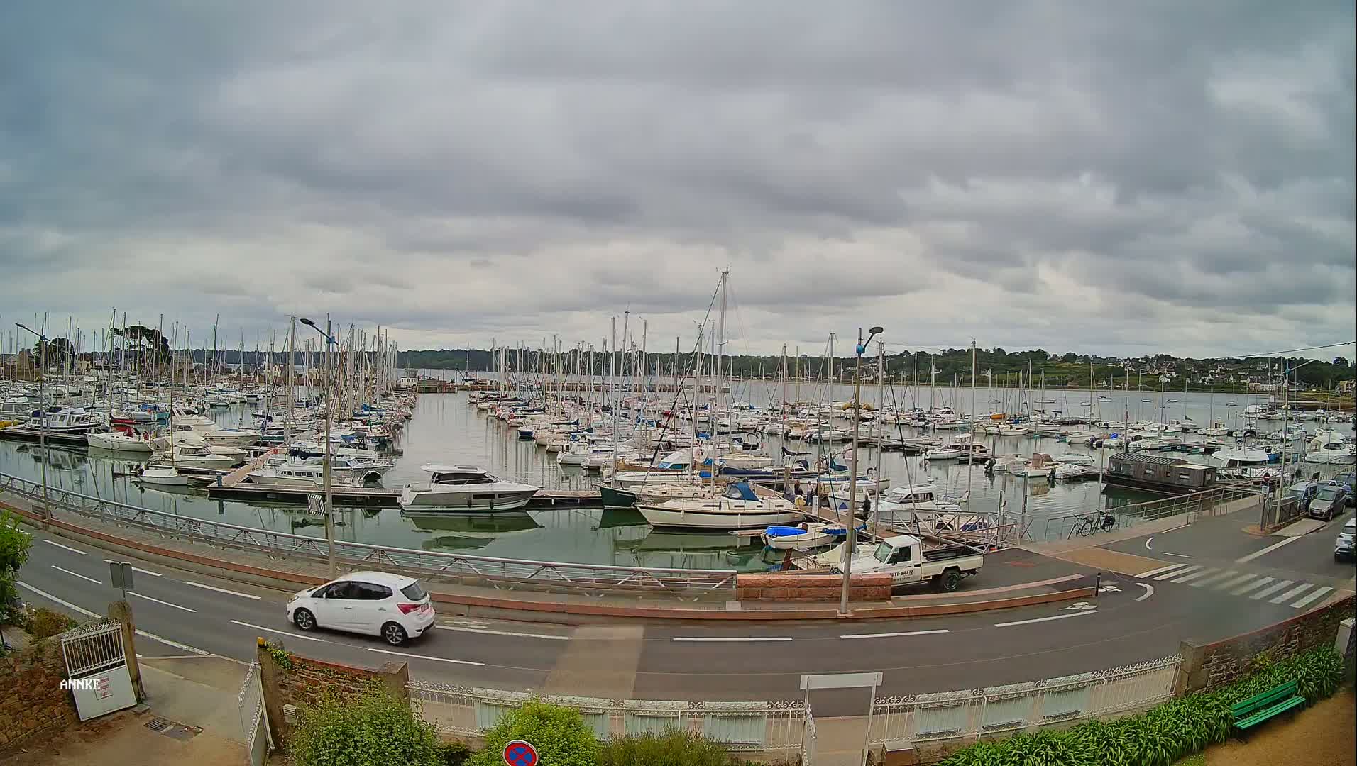 A harbor filled with numerous sailboats and motorboats under a cloudy sky, with a road and a few cars visible in the foreground.