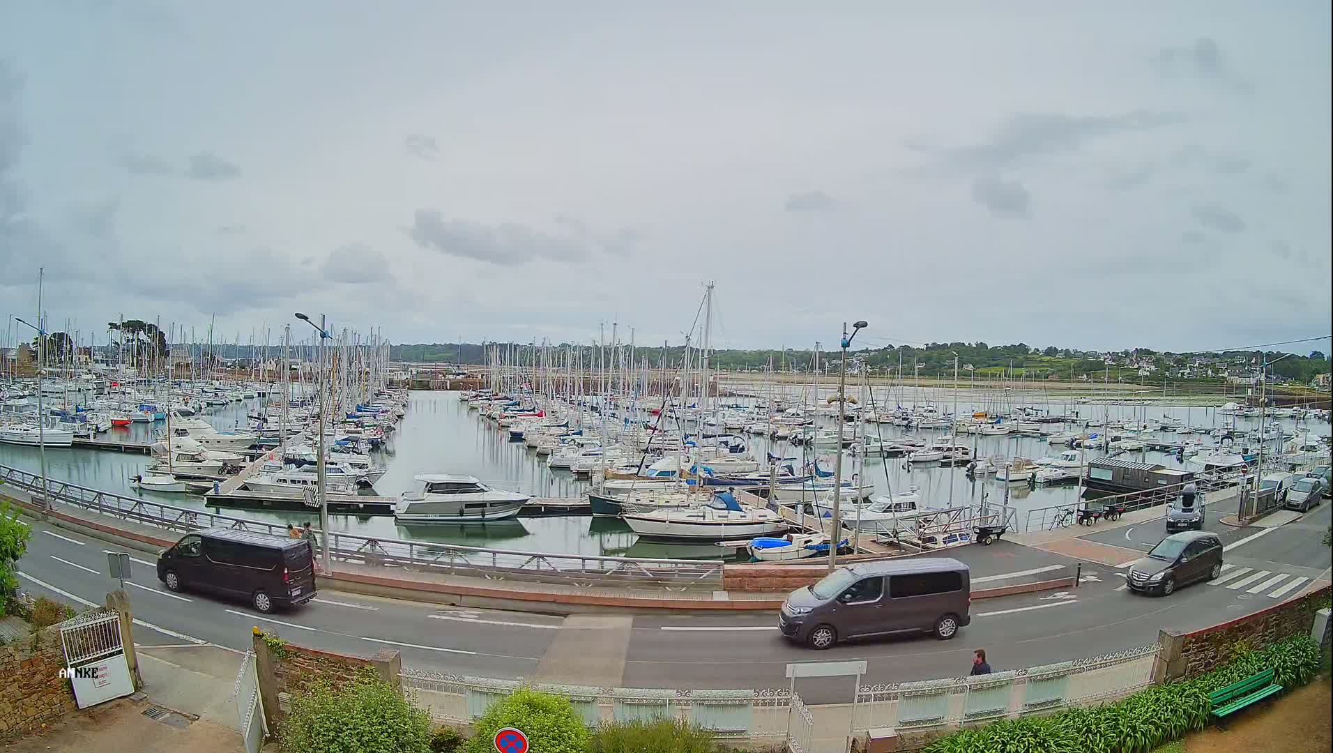 A harbor filled with numerous sailboats and motorboats under a cloudy sky, with a road and several vehicles visible nearby.
