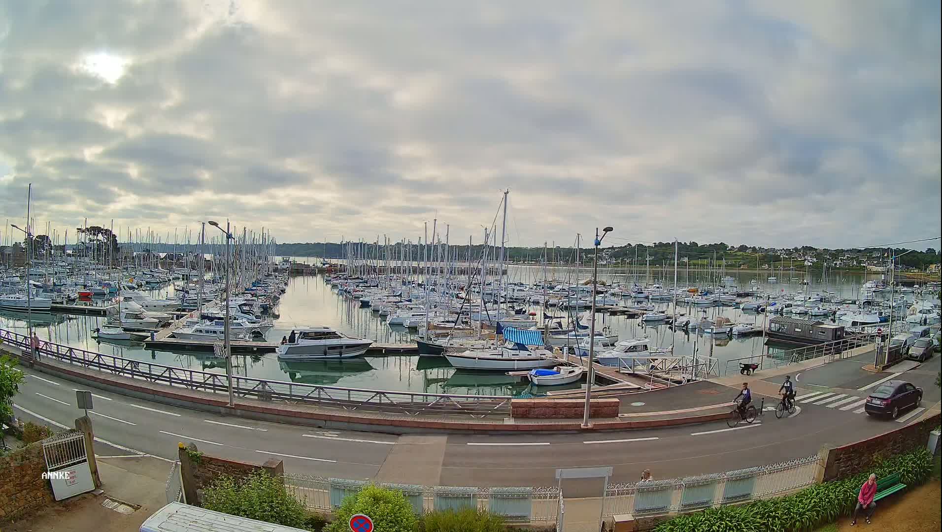 A large marina filled with numerous sailboats and motorboats is seen under a cloudy sky, with a road and cyclists visible in the foreground.