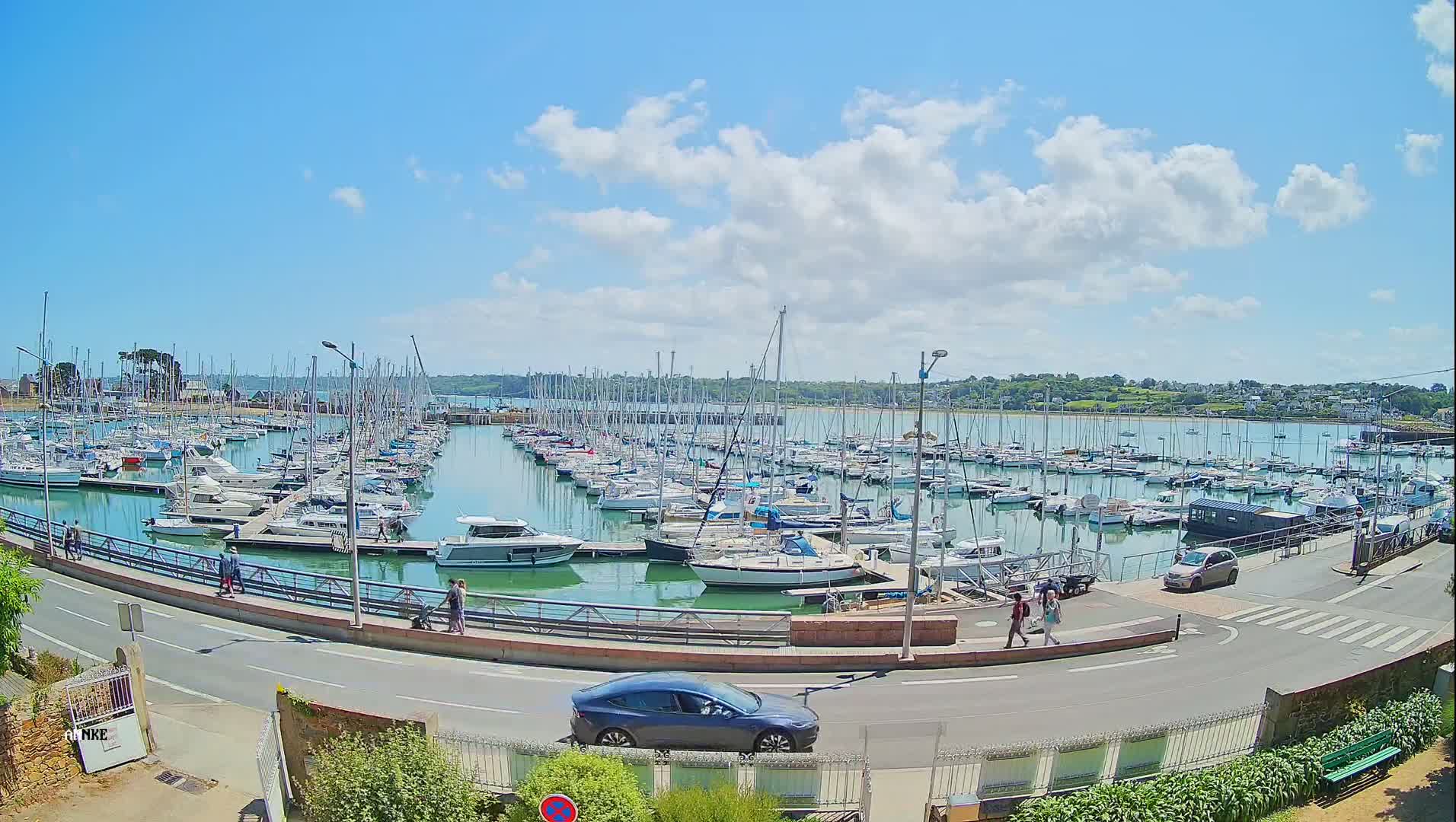 A sunny day with partly cloudy skies shows a marina filled with numerous sailboats and motorboats, a roadway with pedestrians and a car, and green vegetation bordering the scene.