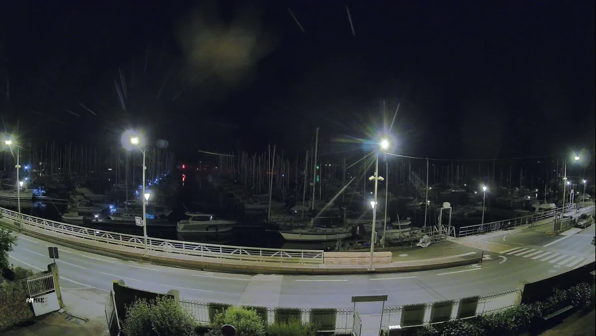 A nighttime, high-angle view of a marina filled with docked sailboats under a dark sky with light streaks.