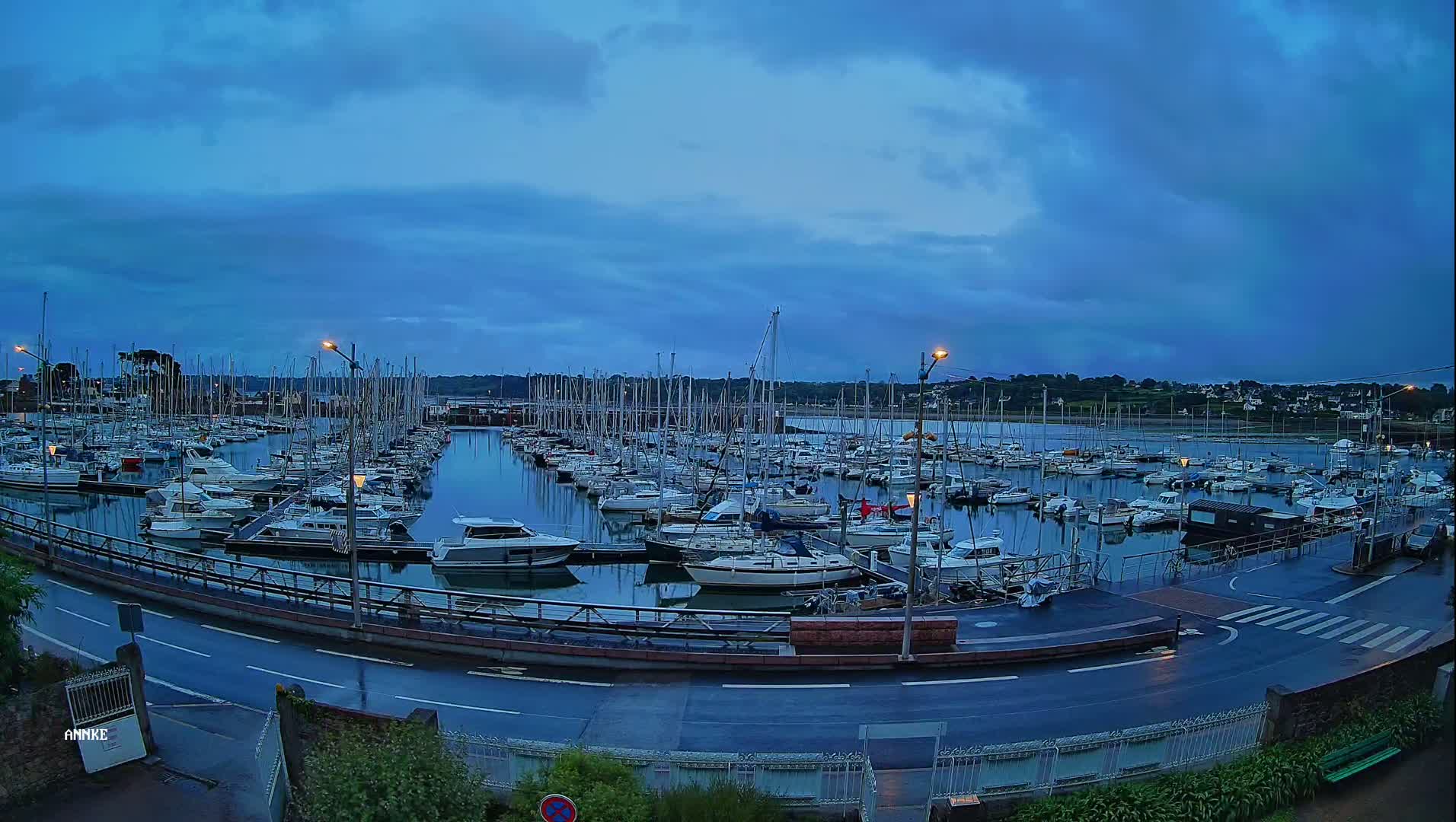 A harbor filled with numerous sailboats is seen under a twilight sky, with a wet road visible in the foreground.