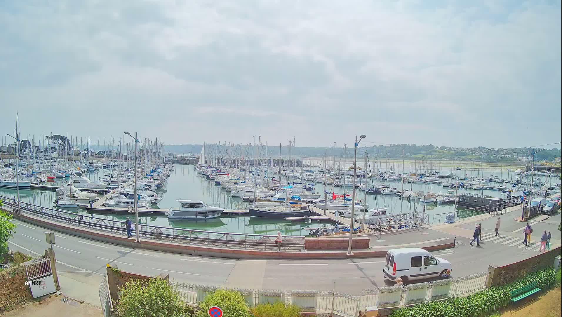 A marina filled with numerous sailboats and motorboats is seen on a partly cloudy day, with a road and pedestrians visible in the foreground.