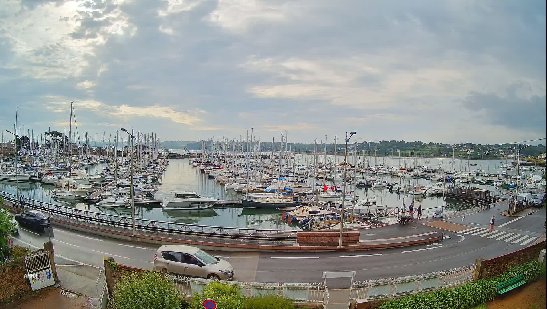 A harbor filled with numerous sailboats and motorboats is seen under a cloudy sky, with a road and a few cars visible in the foreground.