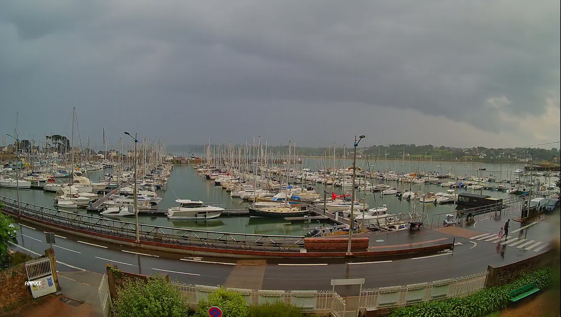 A large marina filled with numerous sailboats and motorboats is shown under a cloudy, rainy sky.