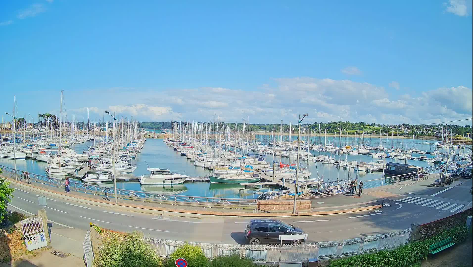 A marina filled with numerous sailboats and motorboats is seen on a partly sunny day, with a road and a few pedestrians visible in the foreground.