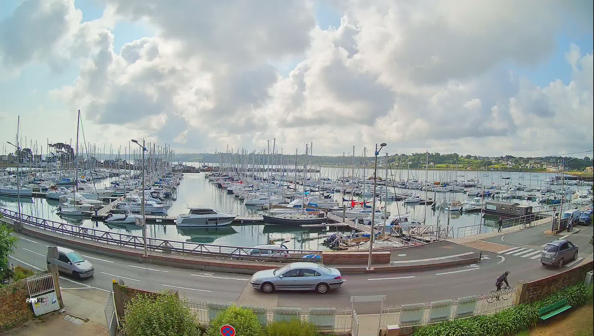 A harbor filled with numerous sailboats and motorboats under a partly cloudy sky, with a road alongside it featuring several cars and a cyclist.