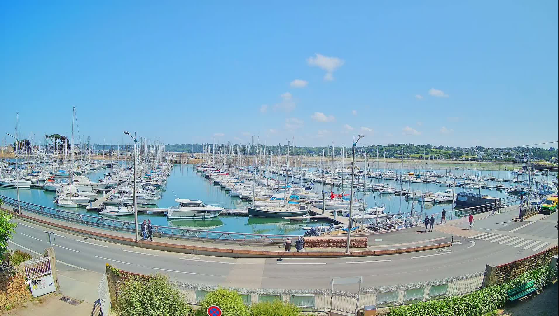 A marina filled with numerous sailboats and motorboats is situated next to a road on a sunny day.