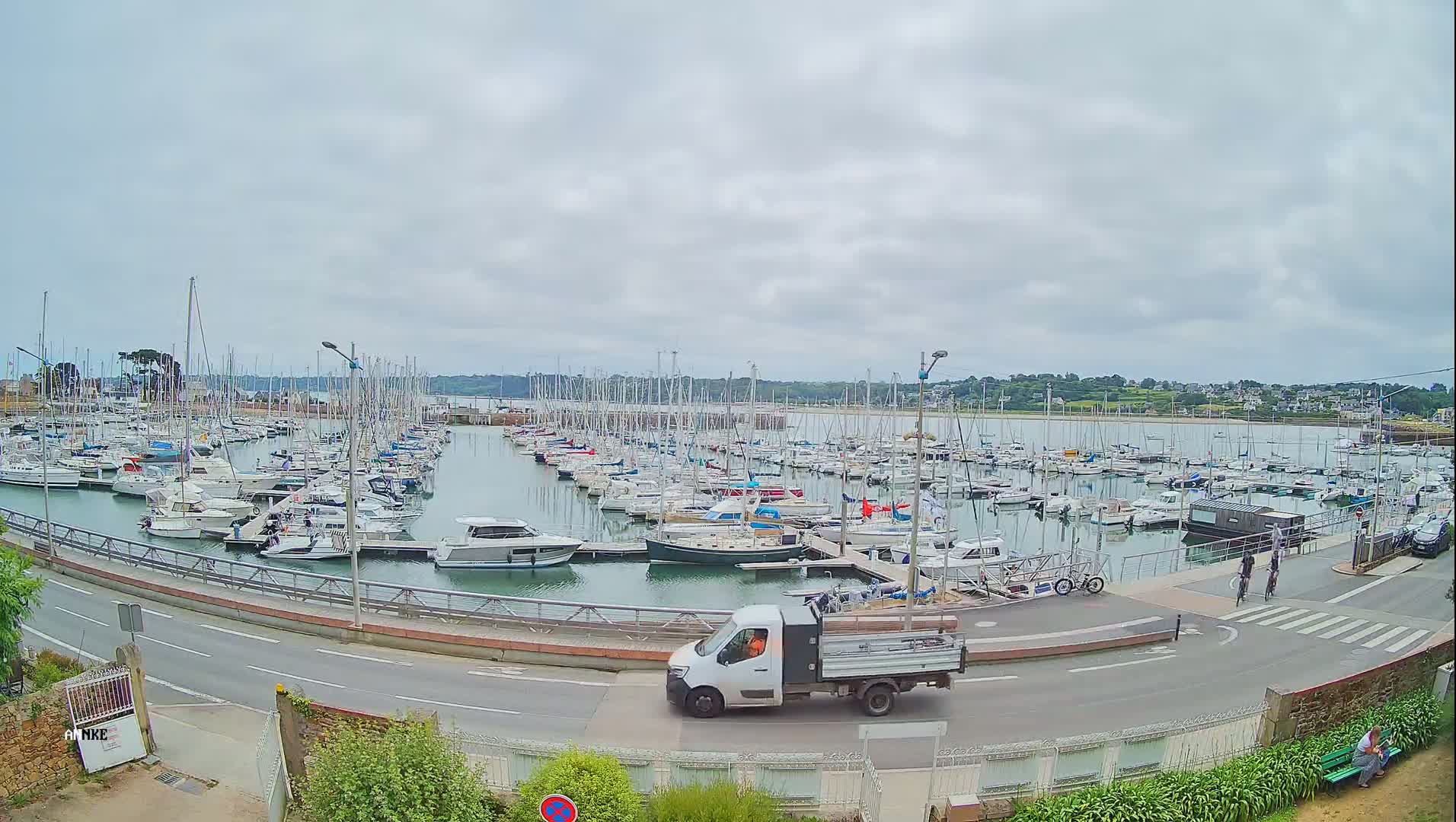 A white pickup truck drives past a marina filled with sailboats and motorboats on an overcast day.
