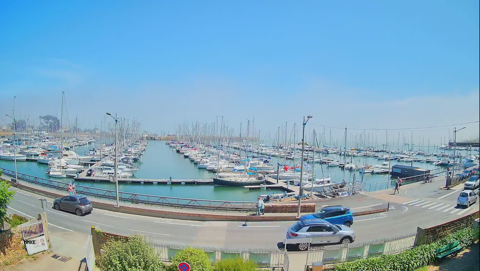 A marina filled with sailboats and motorboats on a sunny day, with cars parked along a nearby road.