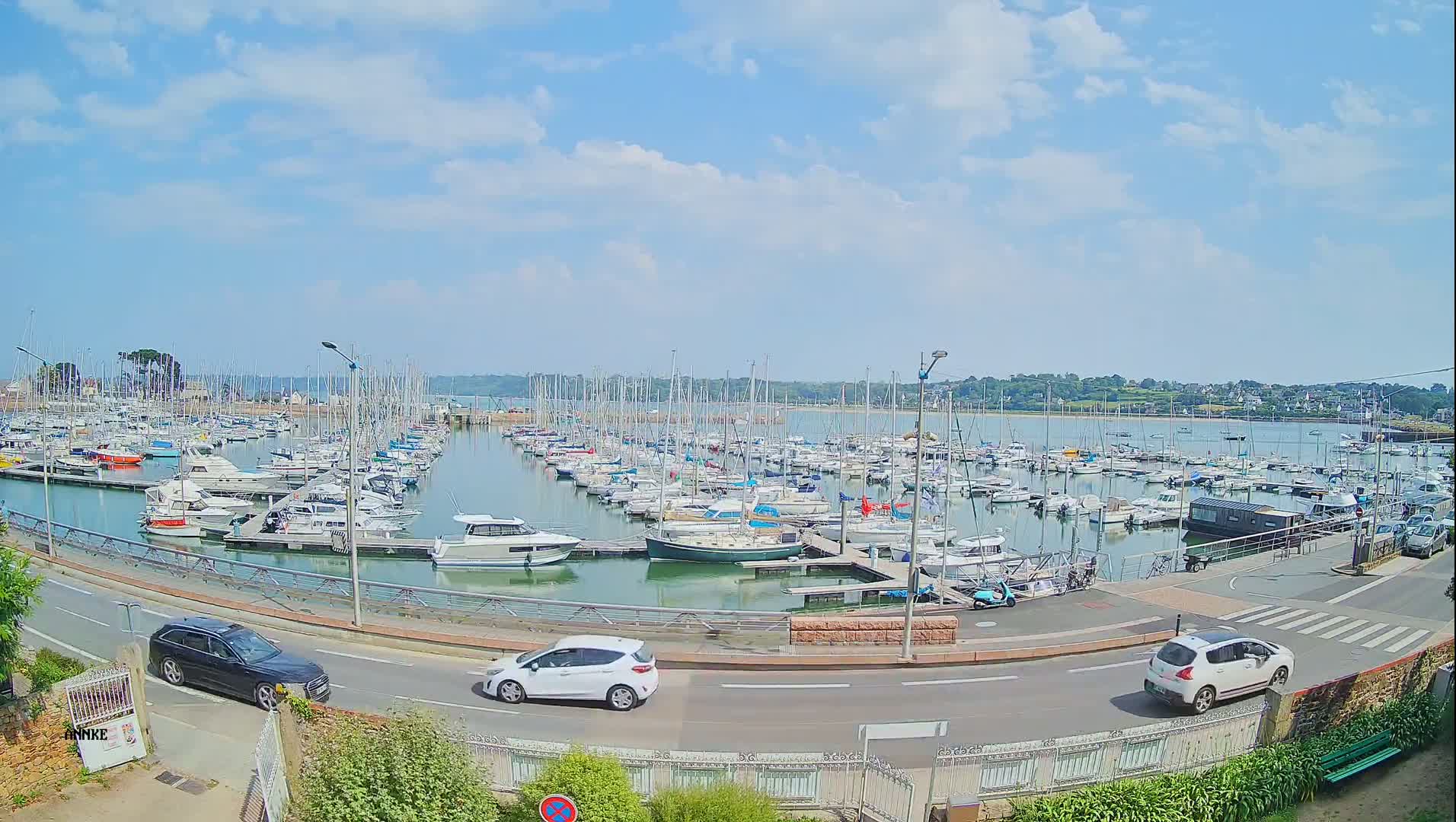 A marina filled with numerous sailboats and motorboats under a mostly sunny sky is viewed from a slightly elevated vantage point, with cars driving on a road in the foreground.