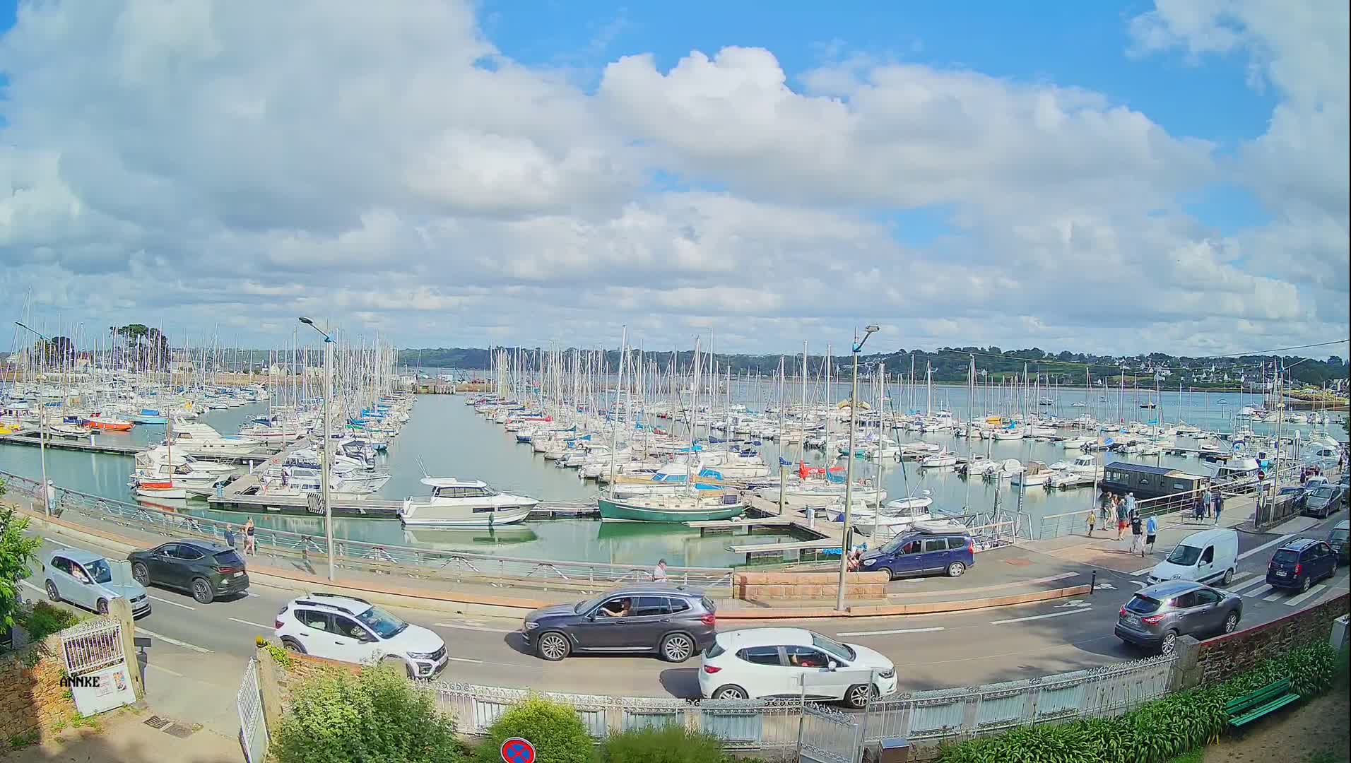 A marina filled with numerous sailboats and motorboats is viewed from a roadside vantage point under a partly cloudy sky.